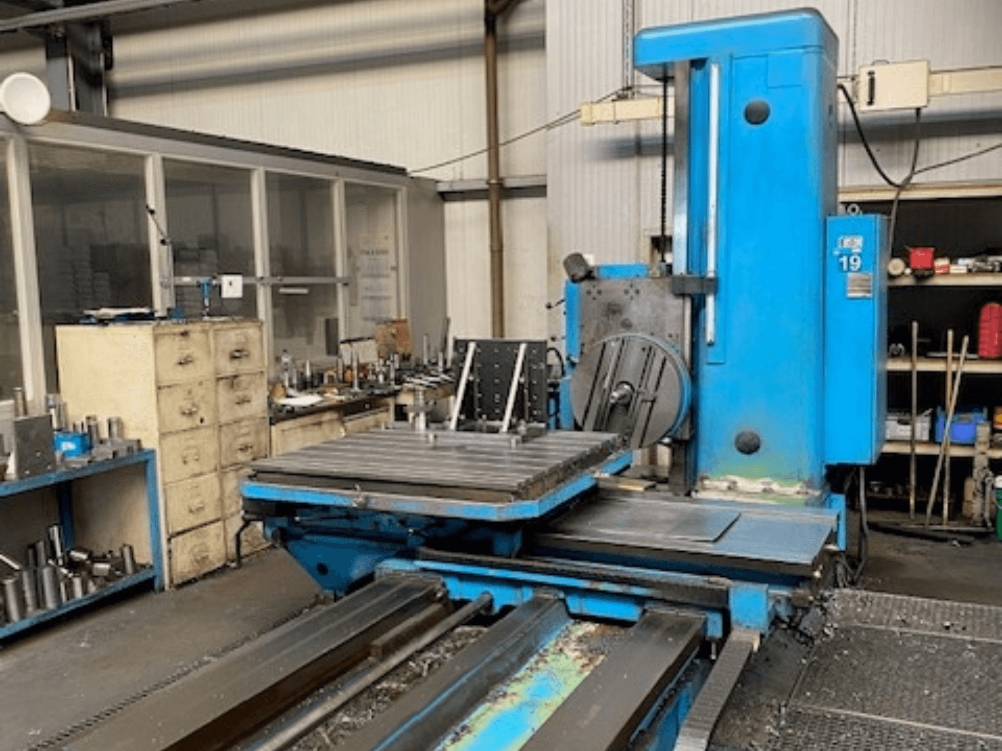 Fresadora azul en vista frontal, modelo número 19, en el suelo de un taller con herramientas y un armario de almacenamiento al fondo.