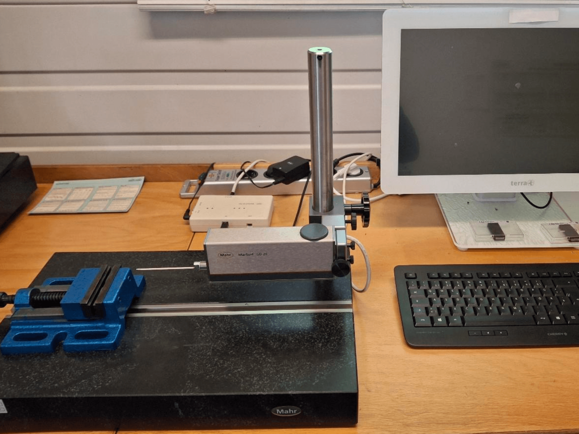MMC MAHR MARSURF GD 26 en un banco de trabajo, con una sonda de medición, una mordaza azul y un monitor de ordenador. Vista frontal.