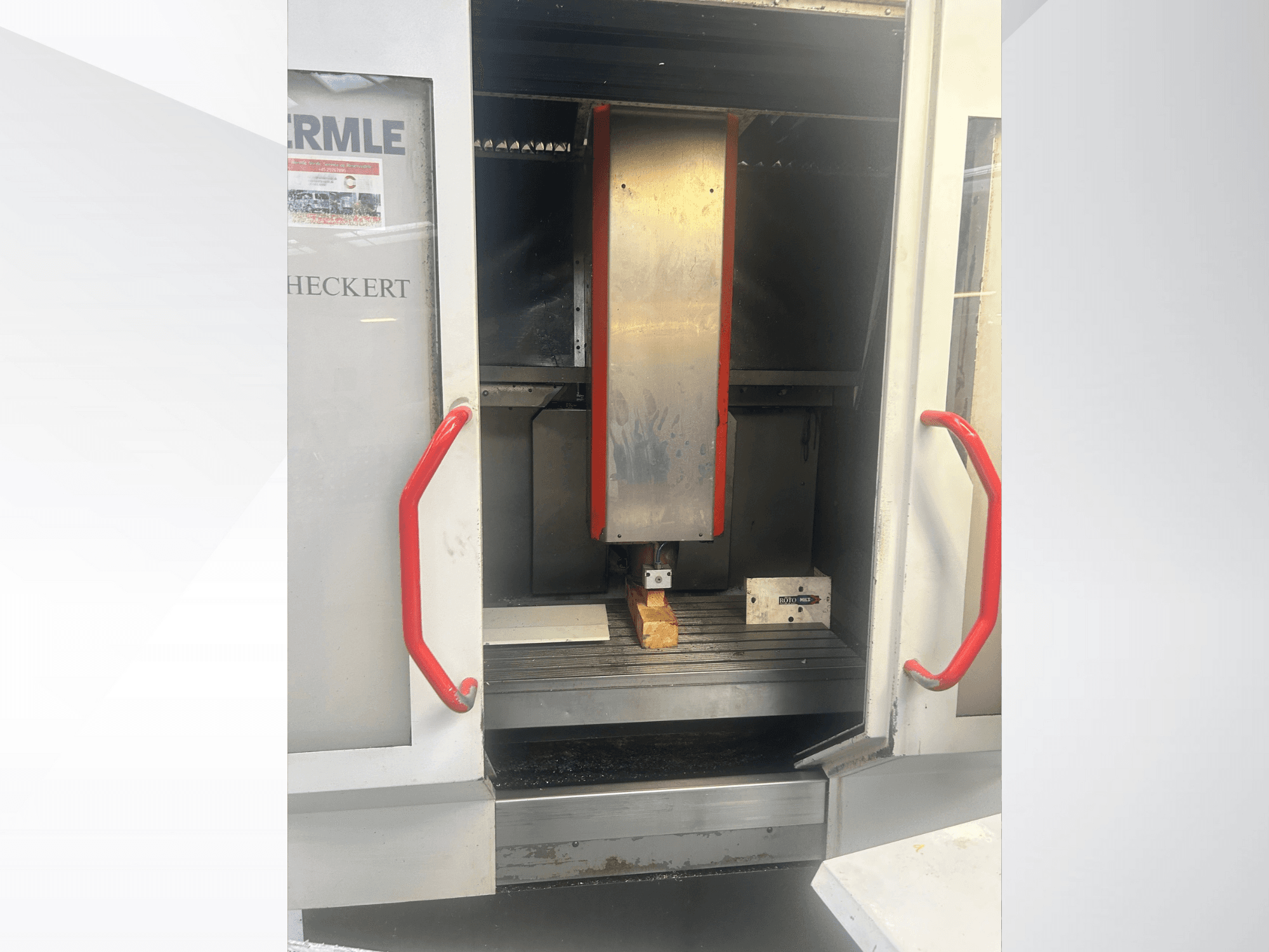 Vista frontal de la máquina CNC de Hermle, con interior metálico, asas rojas y una pieza de trabajo sobre la mesa.