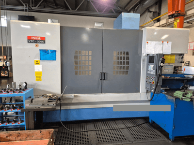 Máquina Mazak VTC-300C en una fábrica, vista frontal mostrando el panel de control, puertas grises con rejilla de ventana y equipos cercanos.