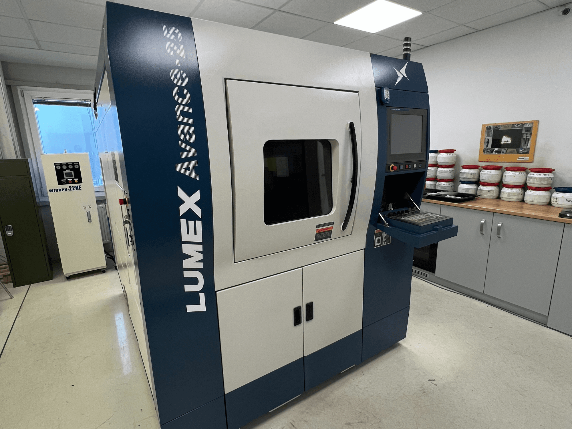 Impresora 3D LUMEX Avance-25 en un laboratorio, vista frontal. Panel de control con pantalla y botones visibles, equipo al fondo.