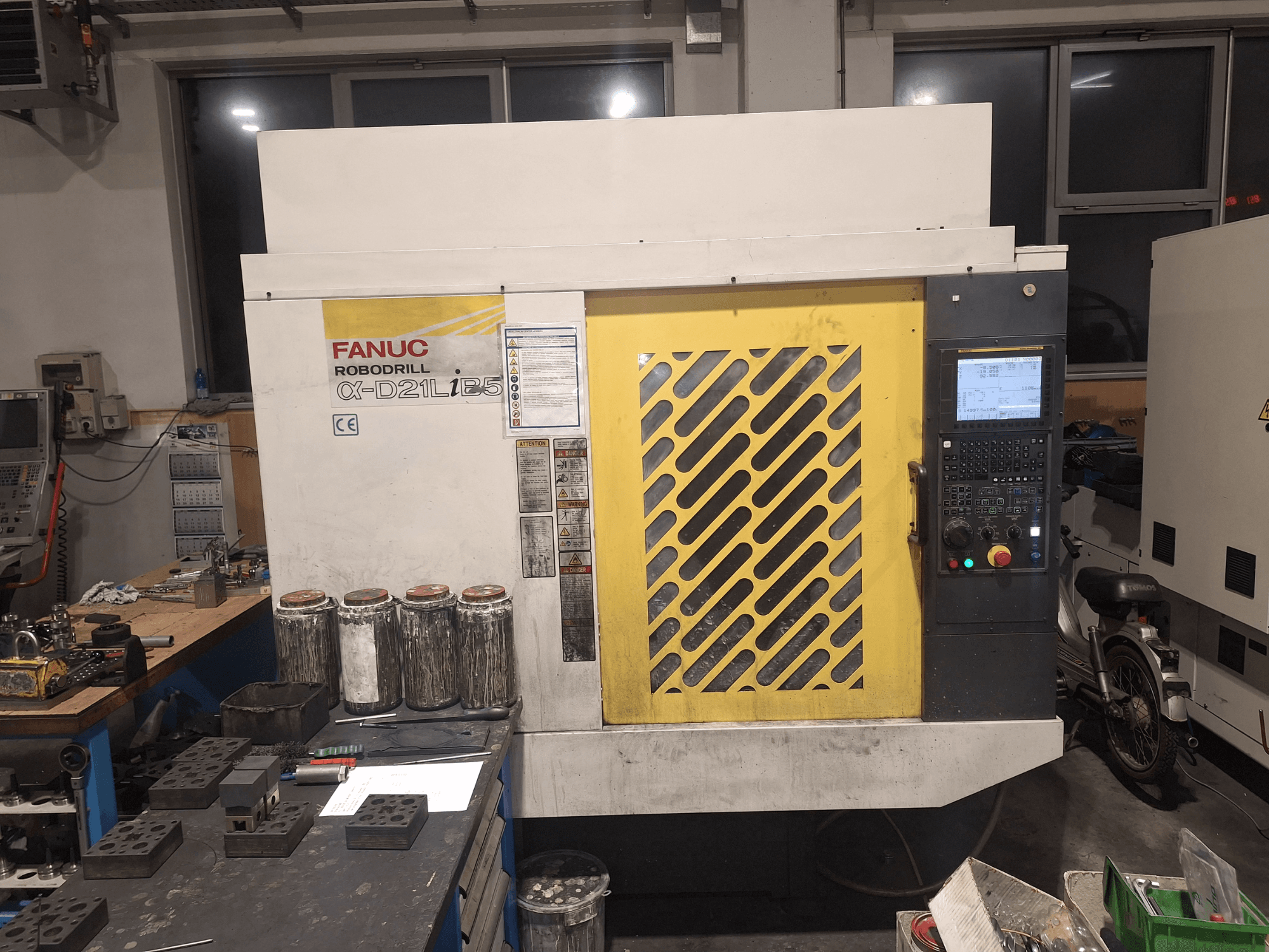 Vista frontal de la FANUC Robodrill α-D21LiB5, mostrando el panel de control, la rejilla amarilla y las herramientas cercanas en un banco de trabajo.
