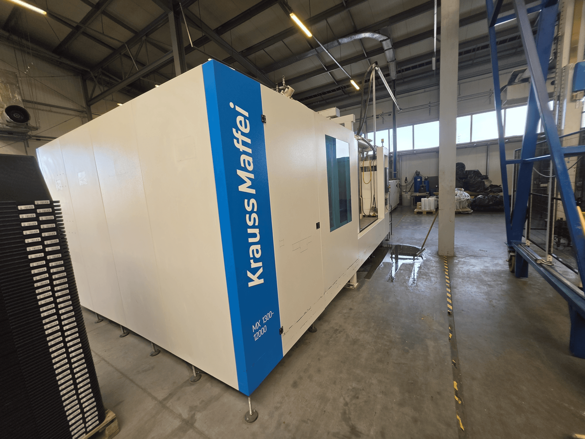 Máquina de moldeo por inyección hidráulica Krauss Maffei MX 1300-12000 vista de perfil, en la que destacan su gran chasis blanco y el logotipo azul de la marca.