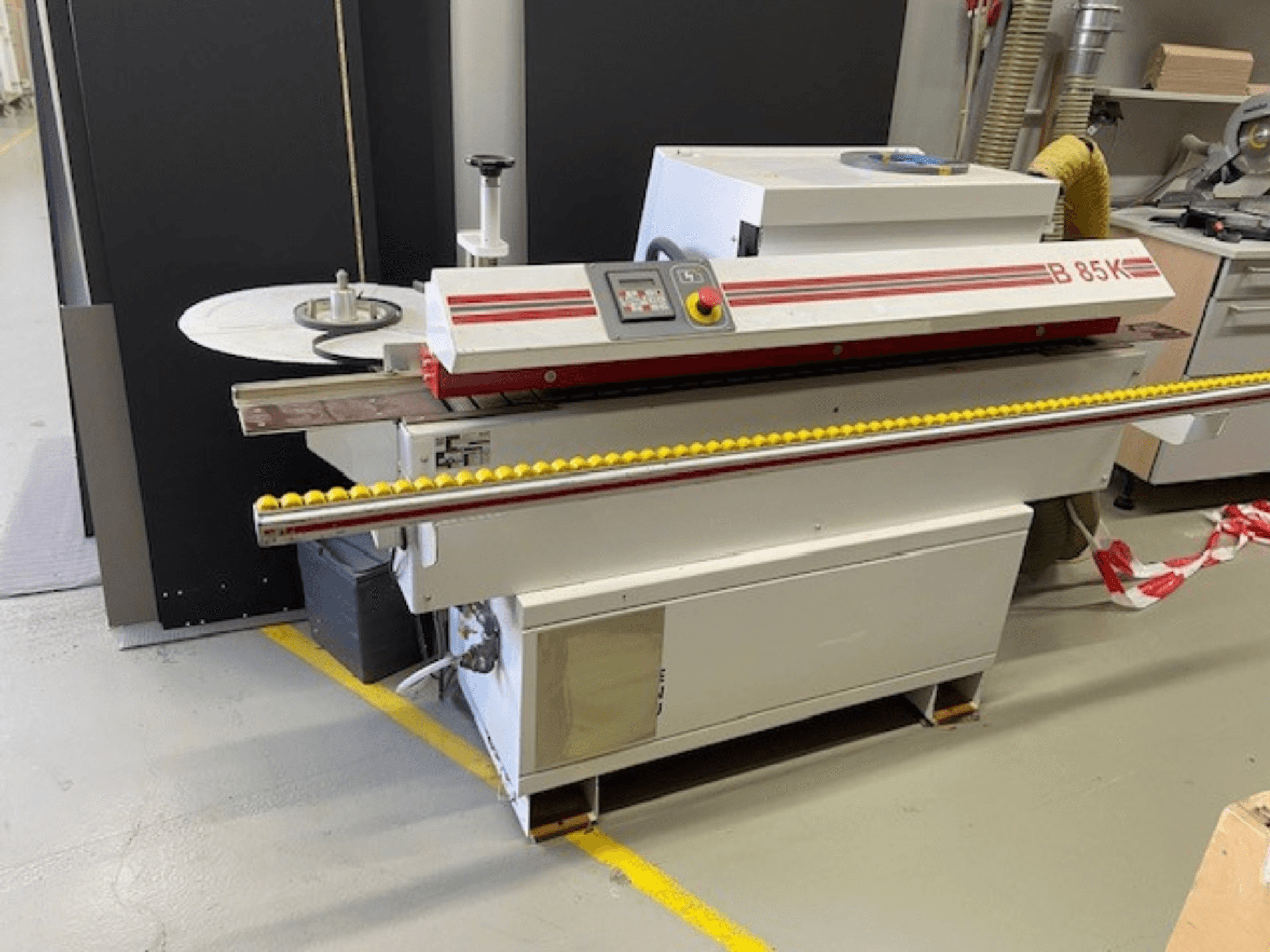 Encoladora de cantos B 85K en blanco y rojo, vista frontal, incorpora un panel de control y un rodillo amarillo para la alimentación del material.