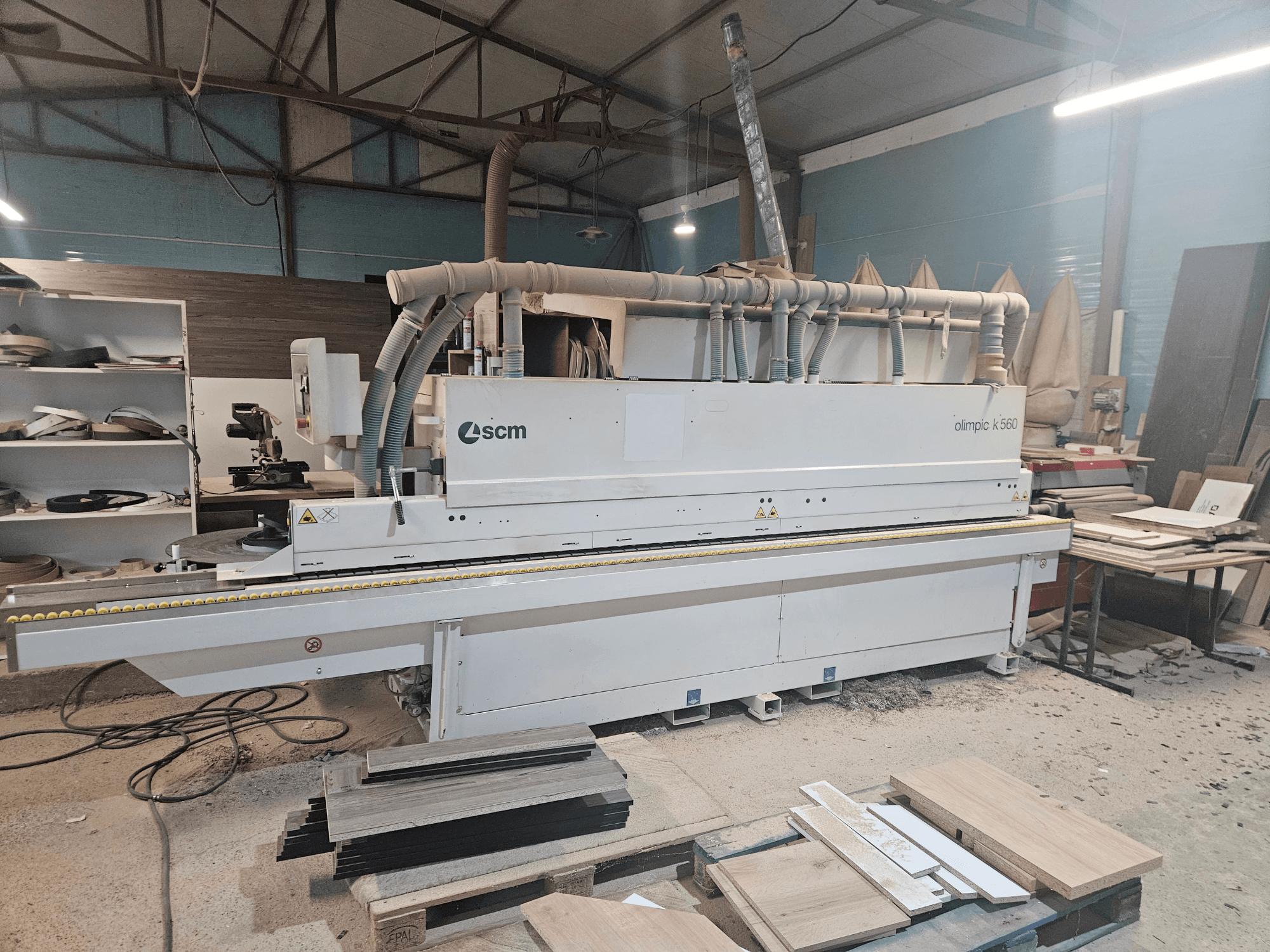 Encoladora de cantos SCM Olimpic K560 en un taller, vista frontal, con paneles de madera y herramientas visibles en el suelo.