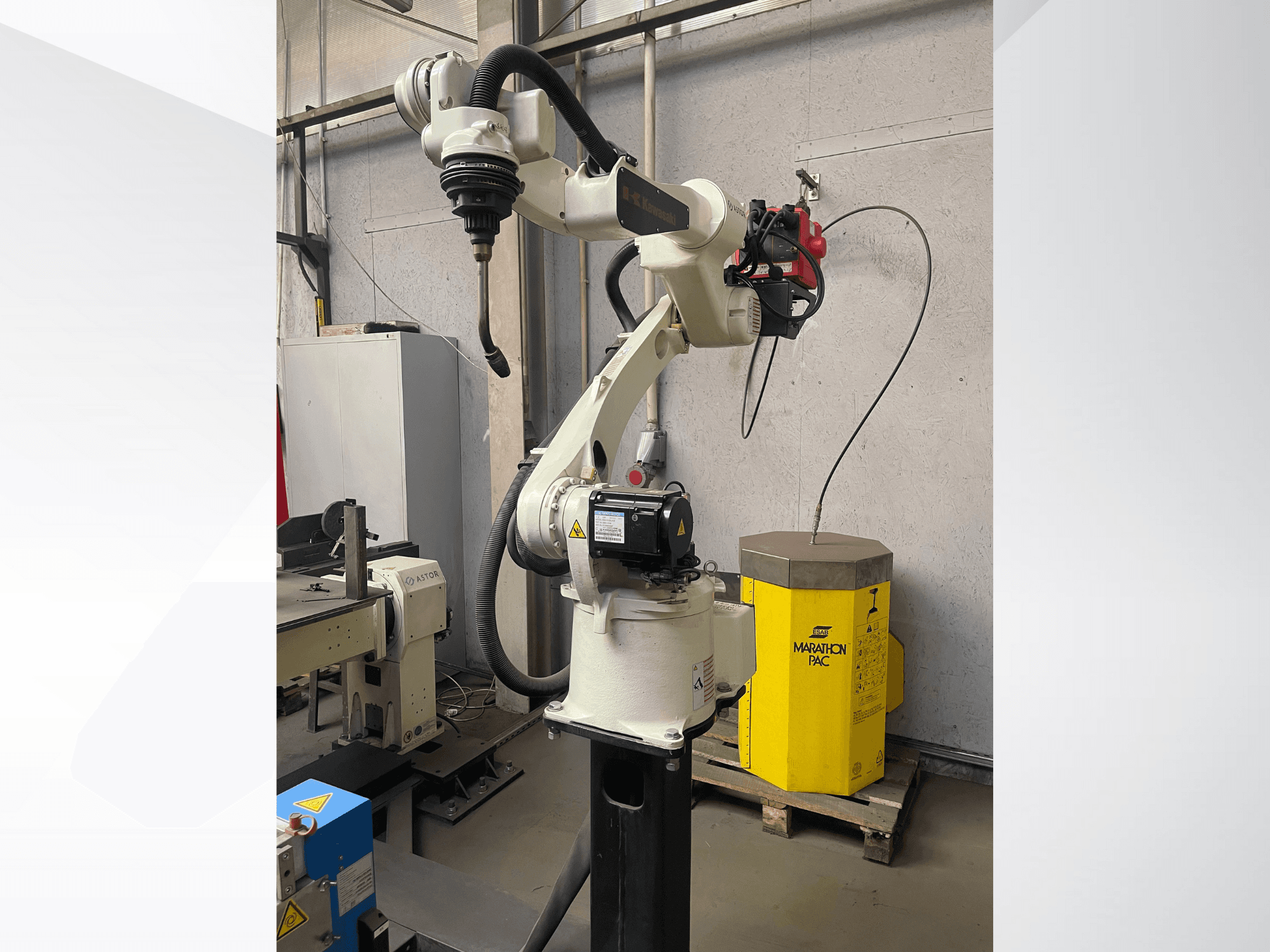 Brazo robótico Kawasaki desde una vista lateral, con un portaherramientas montado y una unidad de potencia amarilla al fondo.