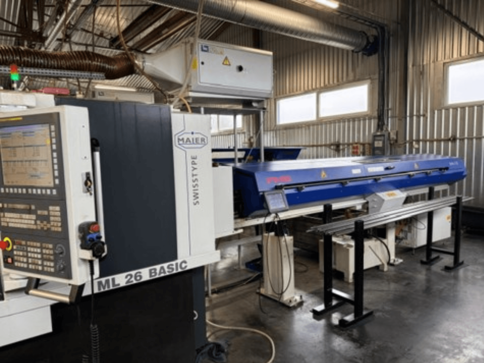 Texto alternativo: Vista frontal de una máquina de torneado Maier ML26 C5Y Swiss Type con panel de control y un dispositivo azul acoplado, en un entorno industrial.