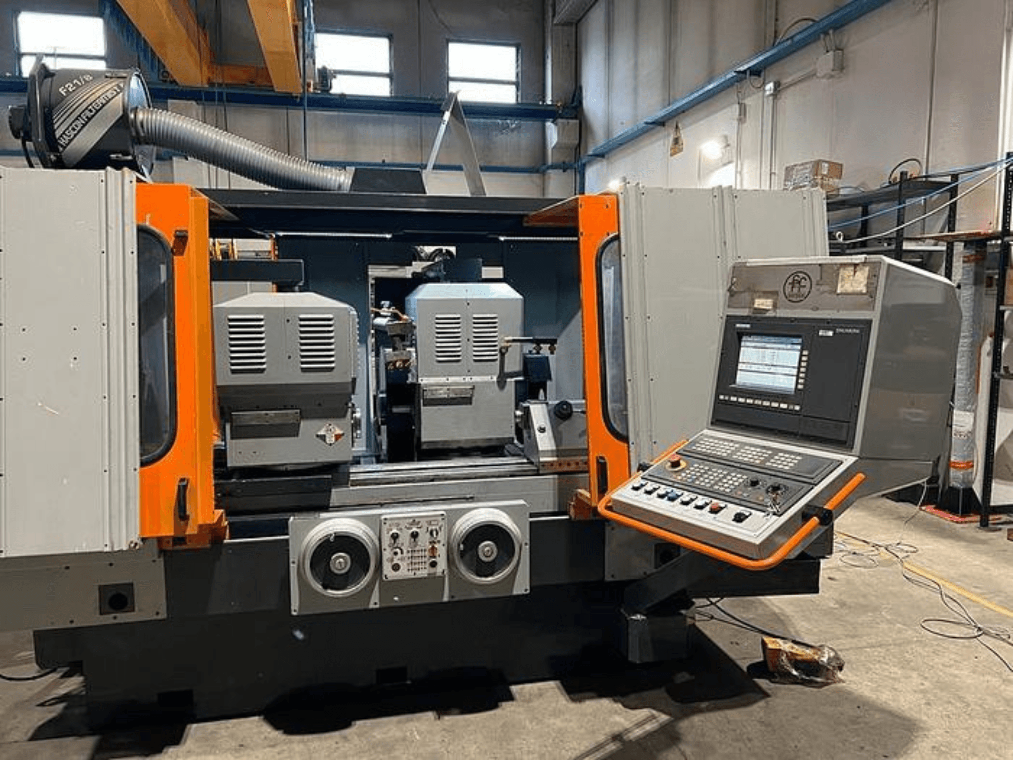 Vista frontal de una máquina CNC con husillos dobles, panel de control y detalles en naranja. Incluye conducto de ventilación y controles de funcionamiento.