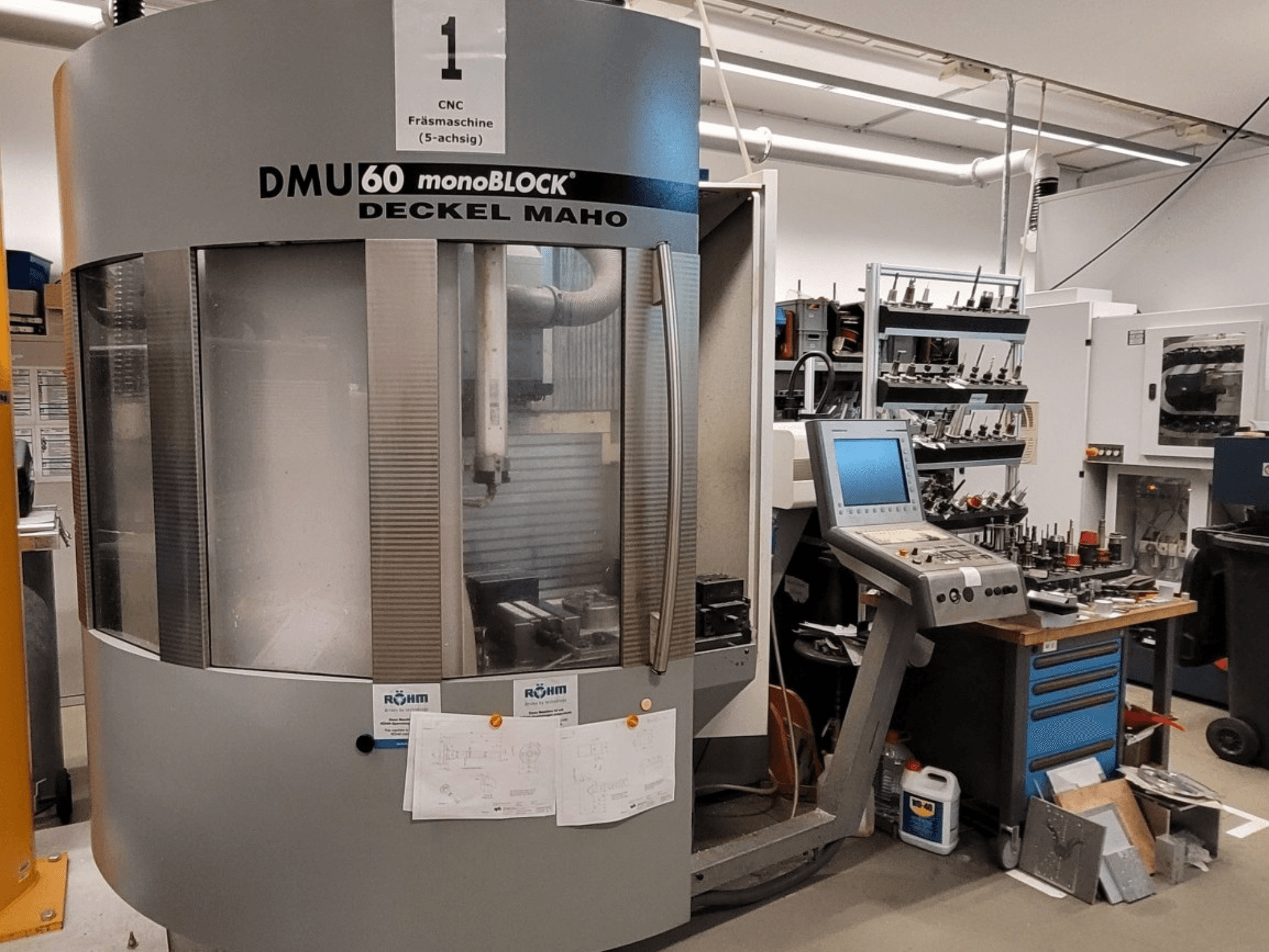 Fresadora CNC DECKEL MAHO DMU 60 monoBLOCK en vista frontal, mostrando el panel de control y el almacén de herramientas junto a ella.