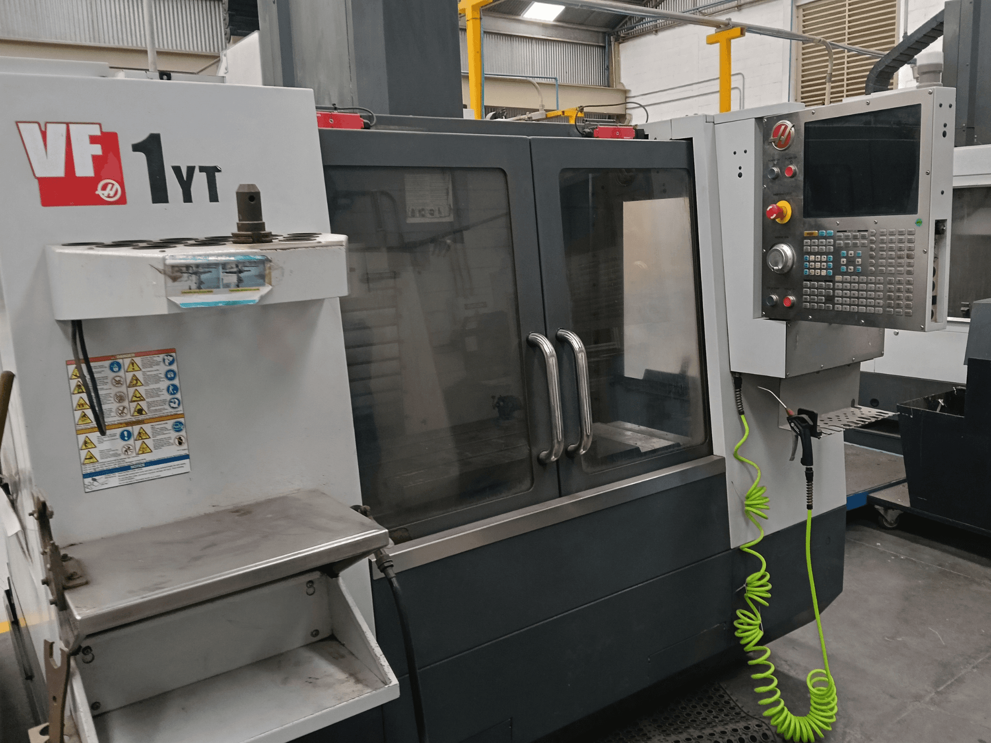Centro de mecanizado vertical HAAS VF-1YT-2, vista frontal, en la que se aprecian el panel de control, el portaherramientas, las etiquetas de seguridad y la zona de mecanizado.