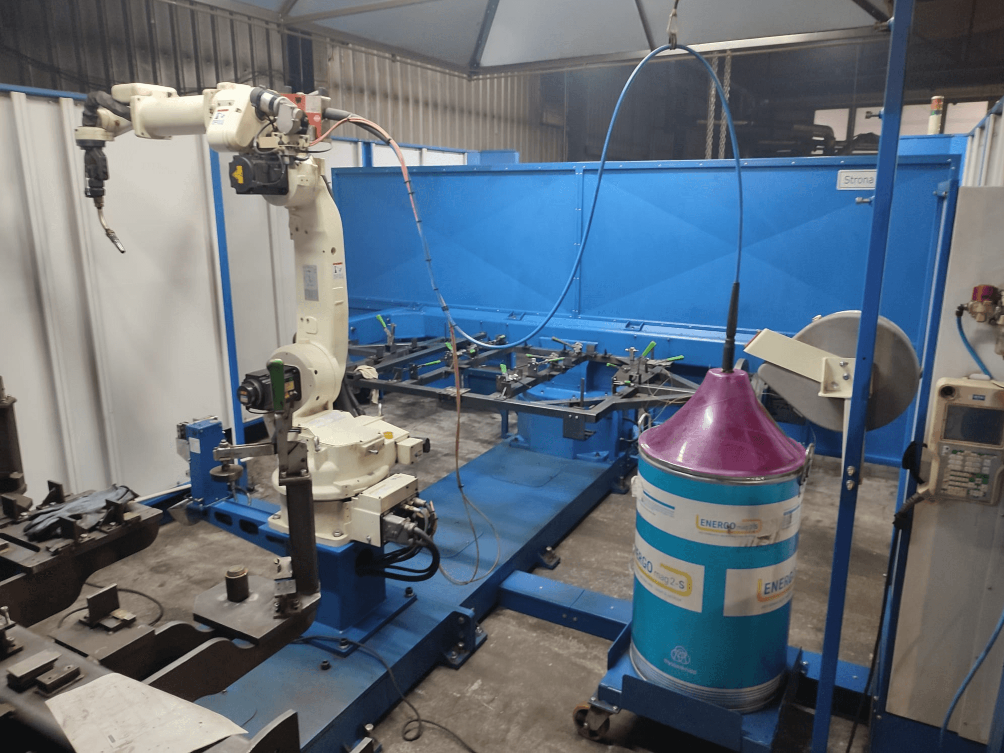 Brazo robótico industrial con base blanca y azul, montado en un taller con una superficie de trabajo azul y componentes metálicos.