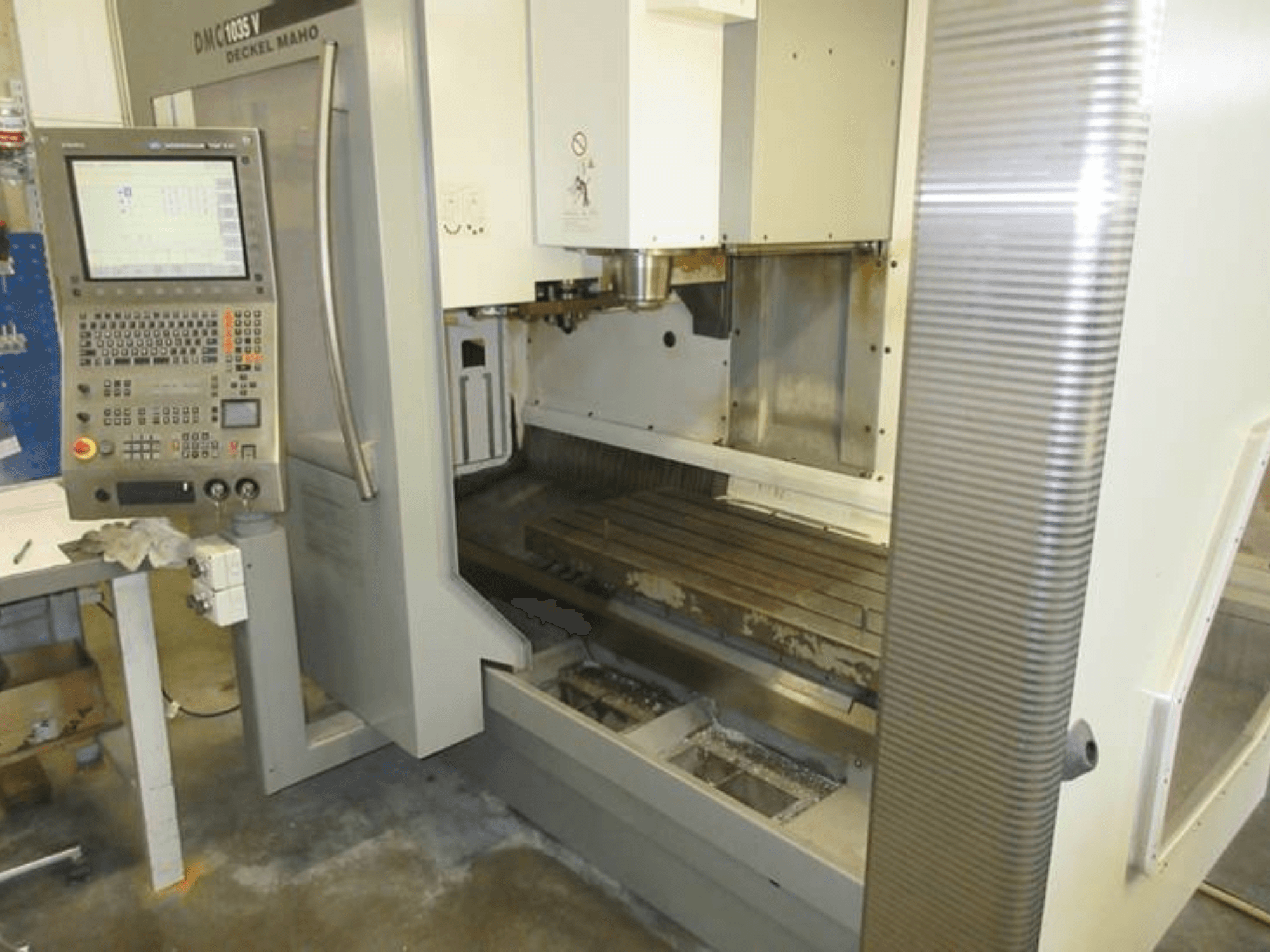 Vista frontal del centro de mecanizado vertical DECKEL MAHO DMC 1035 V, con el panel de control, la mesa de trabajo visible y la zona del husillo.