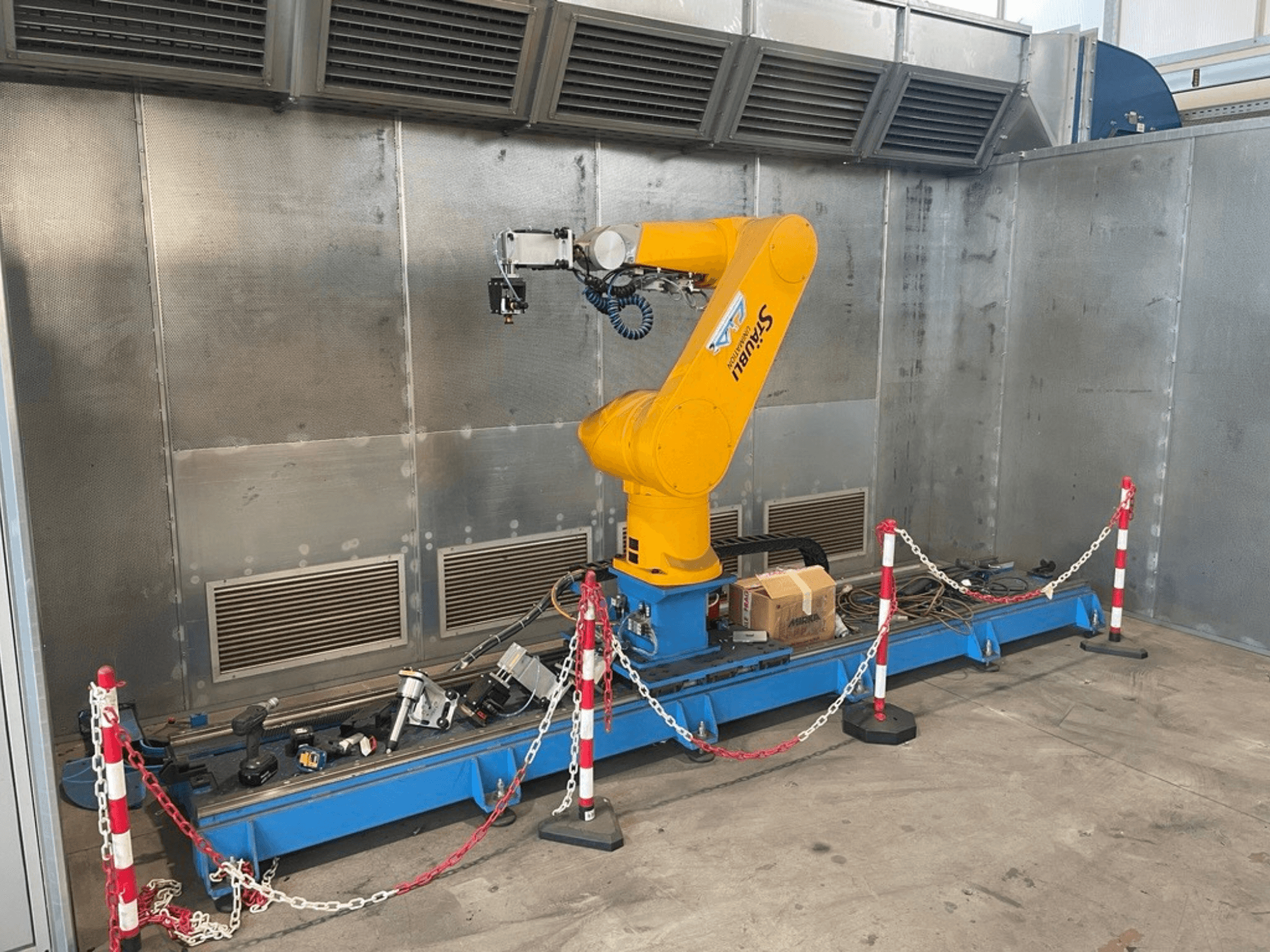 Brazo robótico STAUBLI CS8C-RX160 en un taller, vista lateral, con cables y diversas herramientas sobre una base azul rodeada de barreras protectoras.