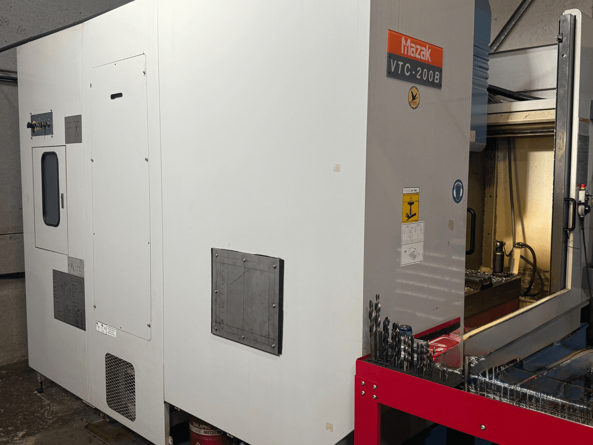 Centro de mecanizado vertical Mazak VTC-200B en vista lateral, en el que se aprecian los mandos etiquetados y las brocas en el estante de almacenamiento rojo.