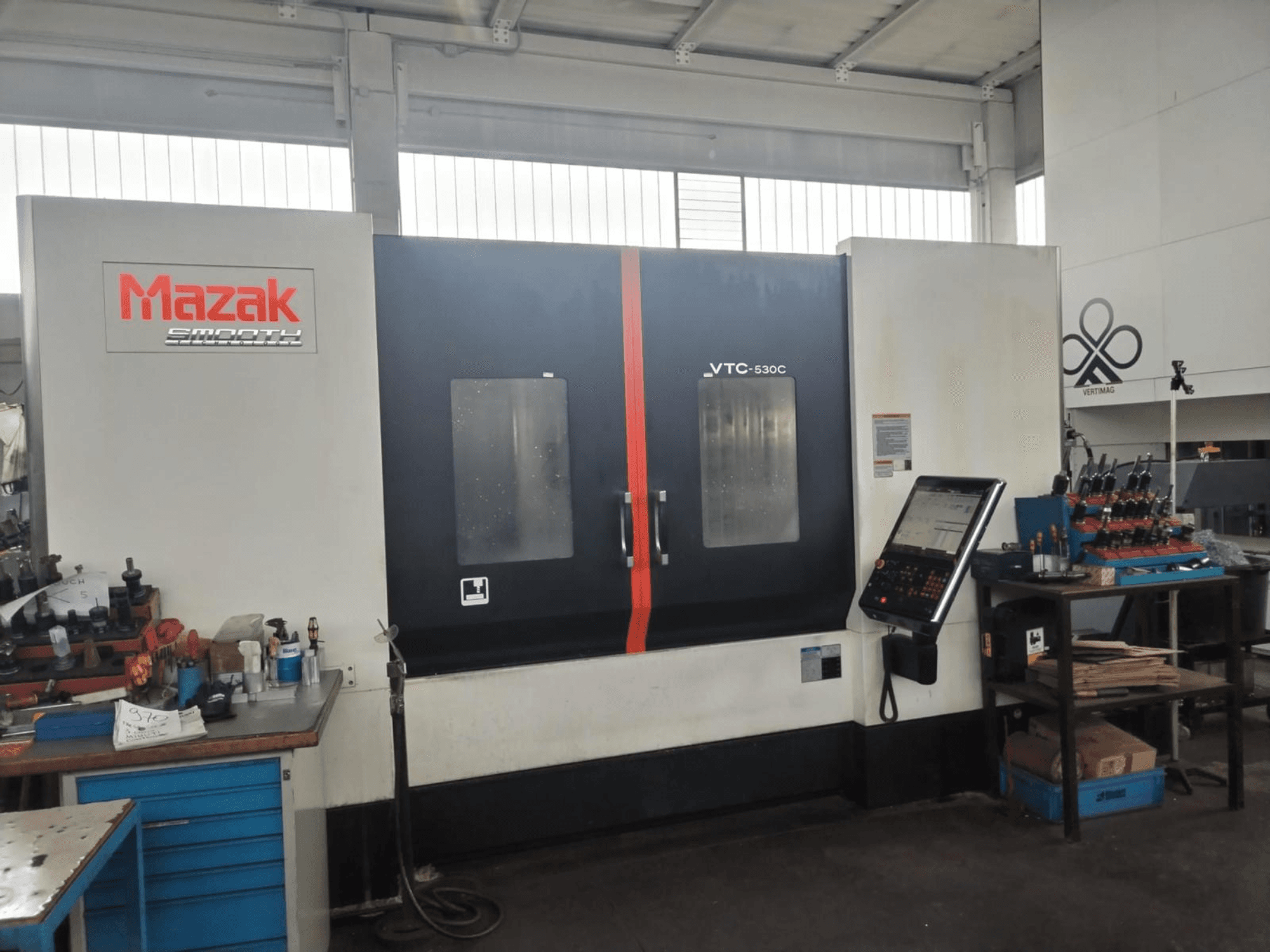 Centro de mecanizado vertical Mazak VTC-530C en un taller, con un panel de control y herramientas en una mesa auxiliar.