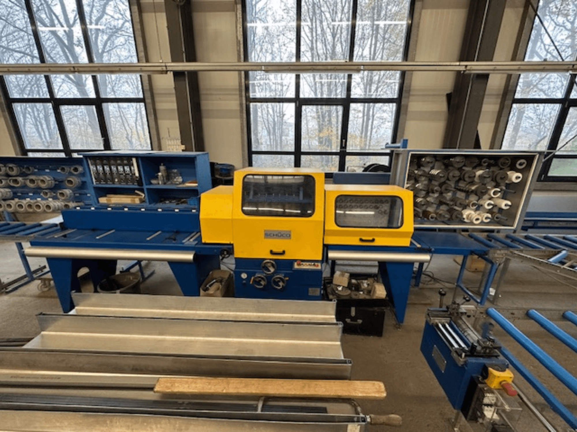 Vista frontal de la máquina herramienta SCHÜCO ISOMAT 4, con un panel de control amarillo, varios rodillos y un compartimento de almacenamiento.