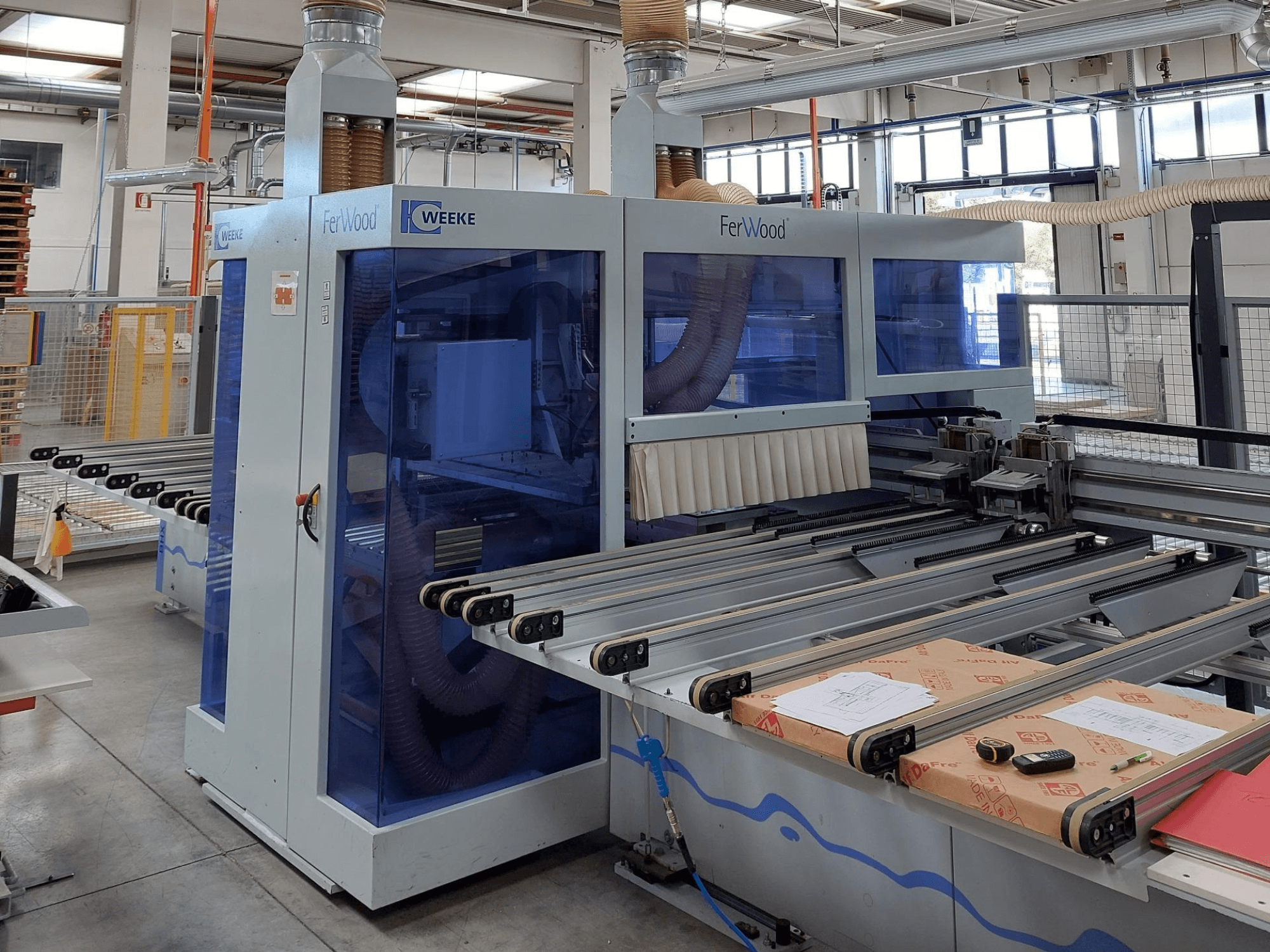 Máquina CNC FerWood de Weeke en un taller, vista frontal que muestra paneles tintados en azul, botones de control y mesa de trabajo con materiales.