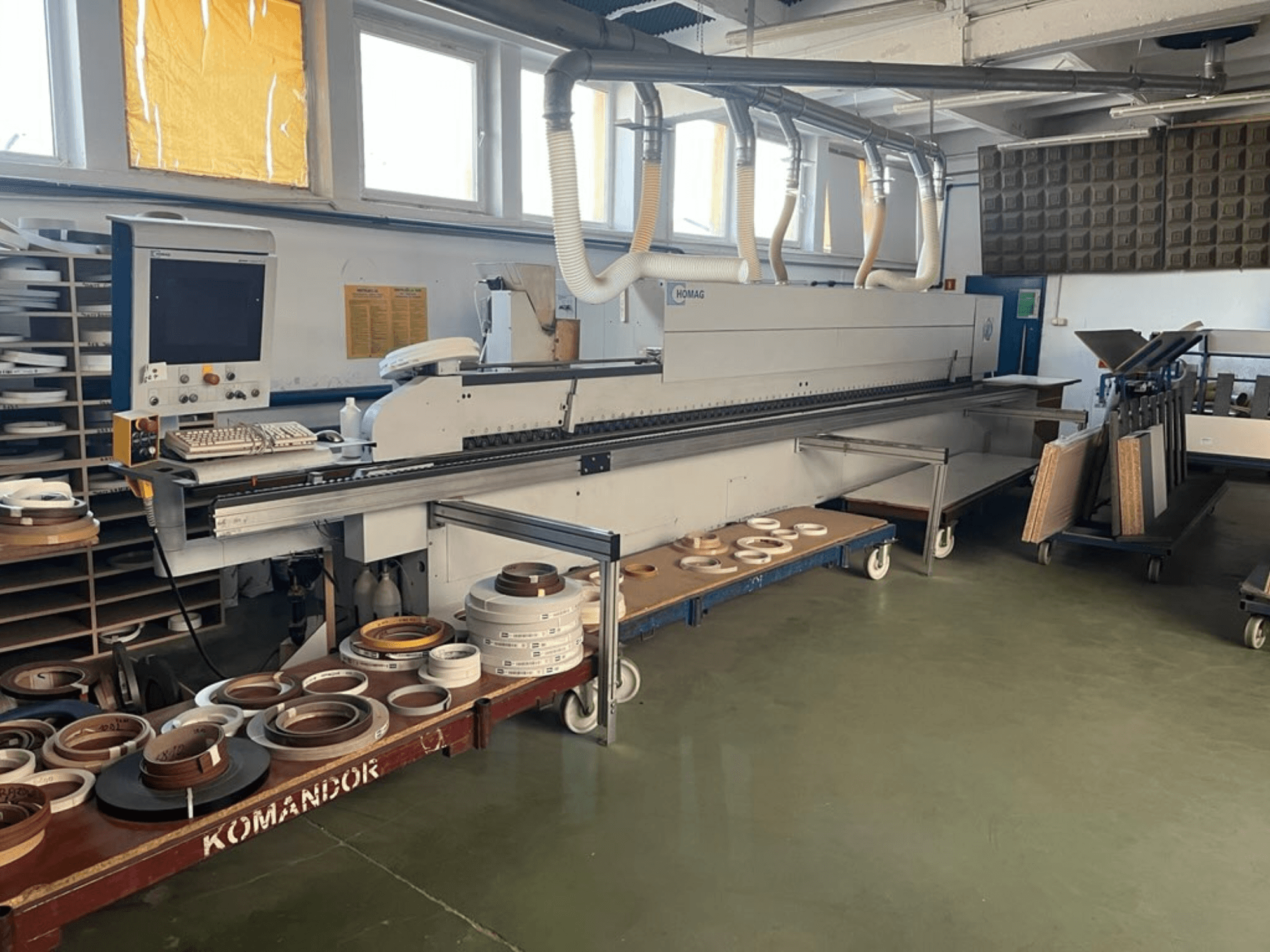 Encoladora de cantos HOMAG en un taller, con controles y diversos materiales de encolado de cantos en un carro azul. Vista lateral.