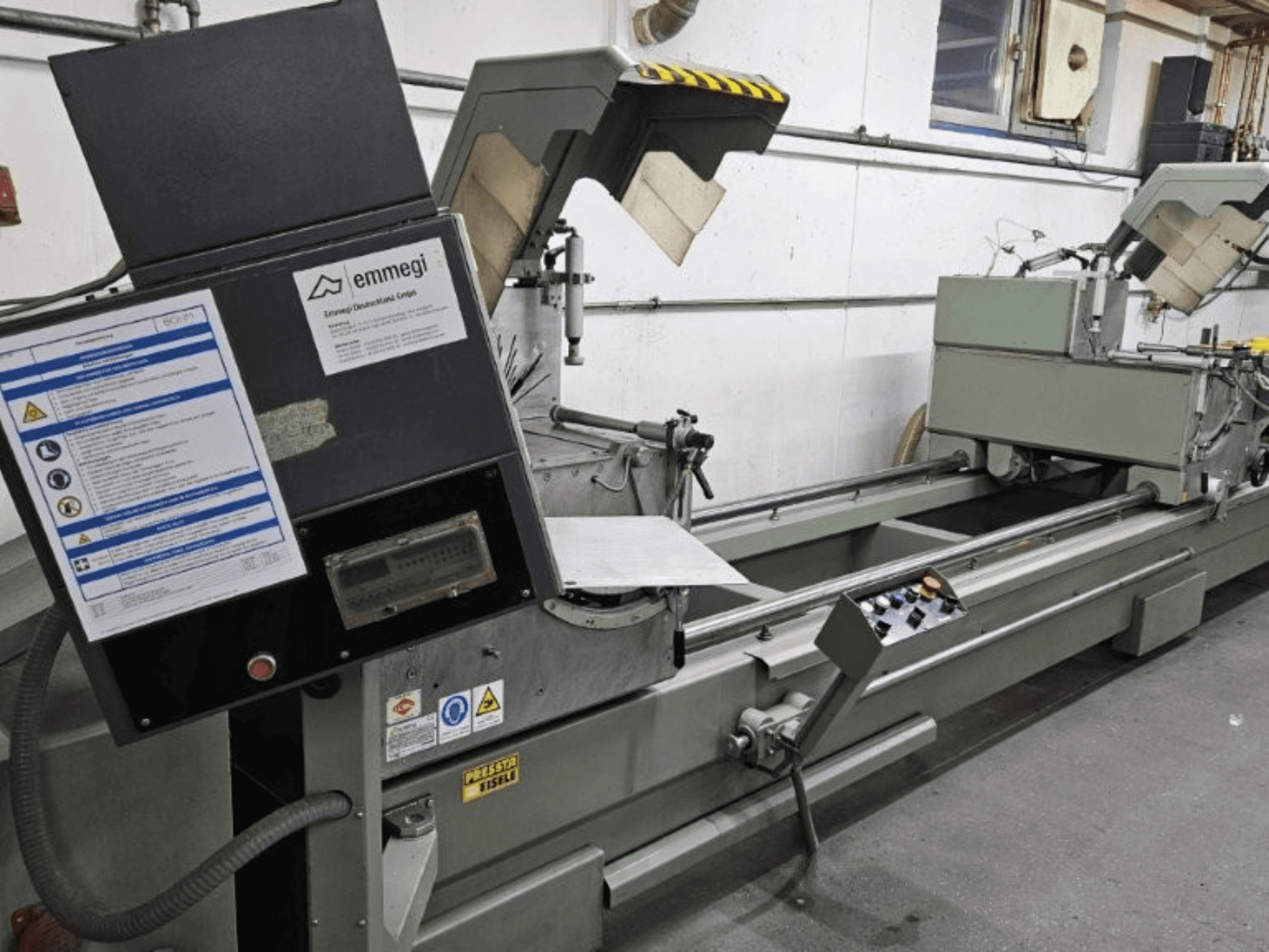 Máquina de corte industrial Emmegi, vista frontal, presenta panel de control, etiquetas de seguridad y componentes operativos.