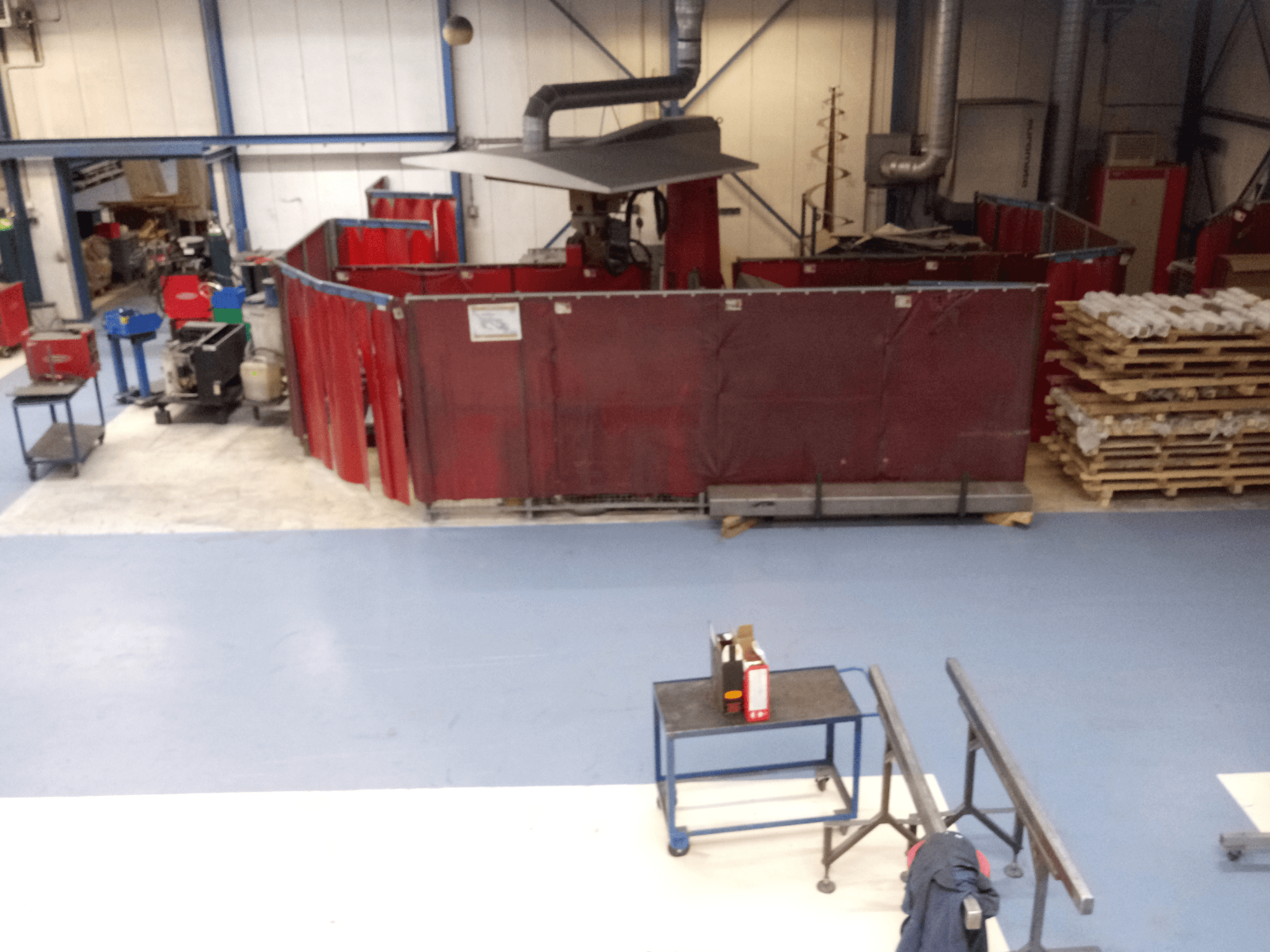 Espacio de trabajo industrial con cortinas de soldadura rojas, máquinas y palés; vista aérea que muestra la disposición y el equipo.