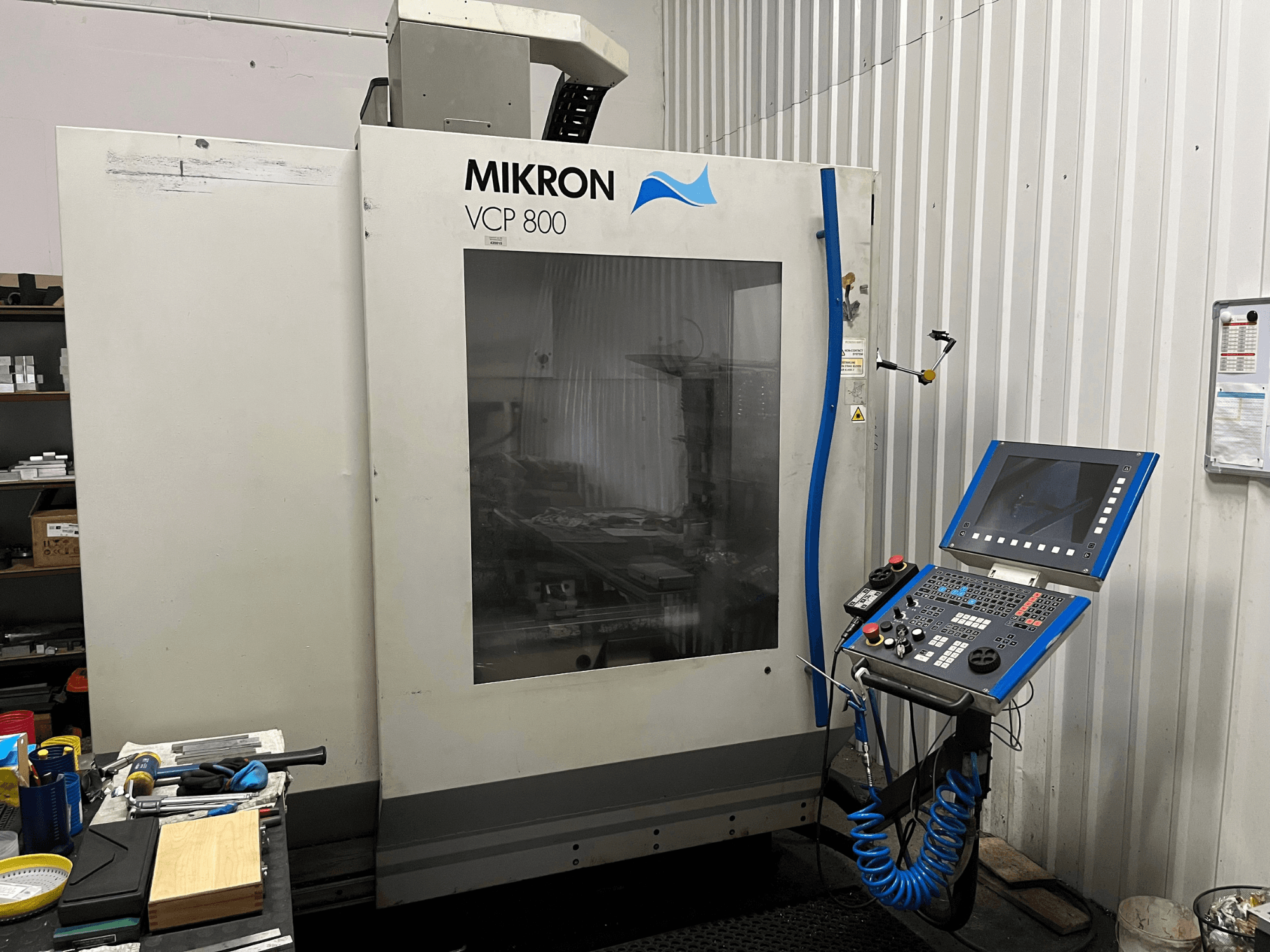Centro de mecanizado vertical MIKRON VCP 800, vista frontal, con el panel de control y las herramientas sobre un banco de trabajo en primer plano.