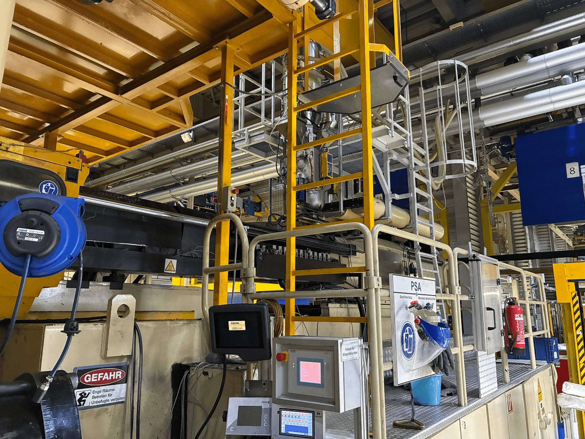 Panel de control de maquinaria industrial con equipo de seguridad azul y andamiaje amarillo, vista lateral mostrando tuberías y señales de advertencia.