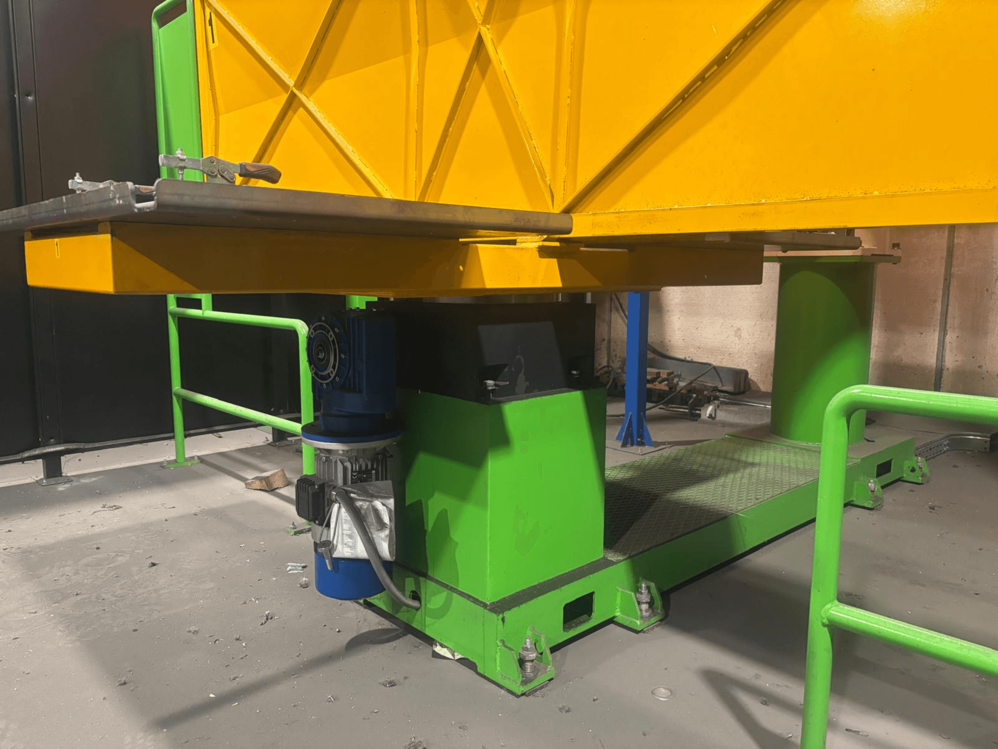 Maquinaria industrial verde y amarilla desde una vista frontal baja, con un motor y una plataforma con una superficie texturizada.