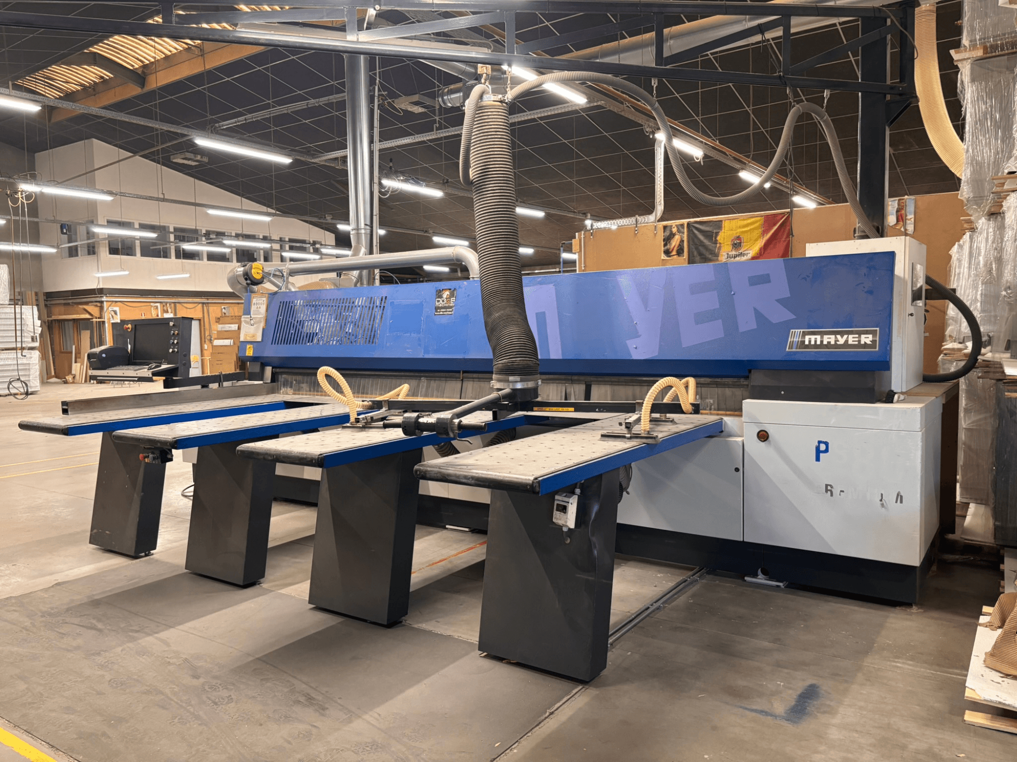 Máquina CNC Mayer con paneles azules, vista frontal; dispone de varias mesas de trabajo y un sistema de aspiración de polvo para trabajar la madera.
