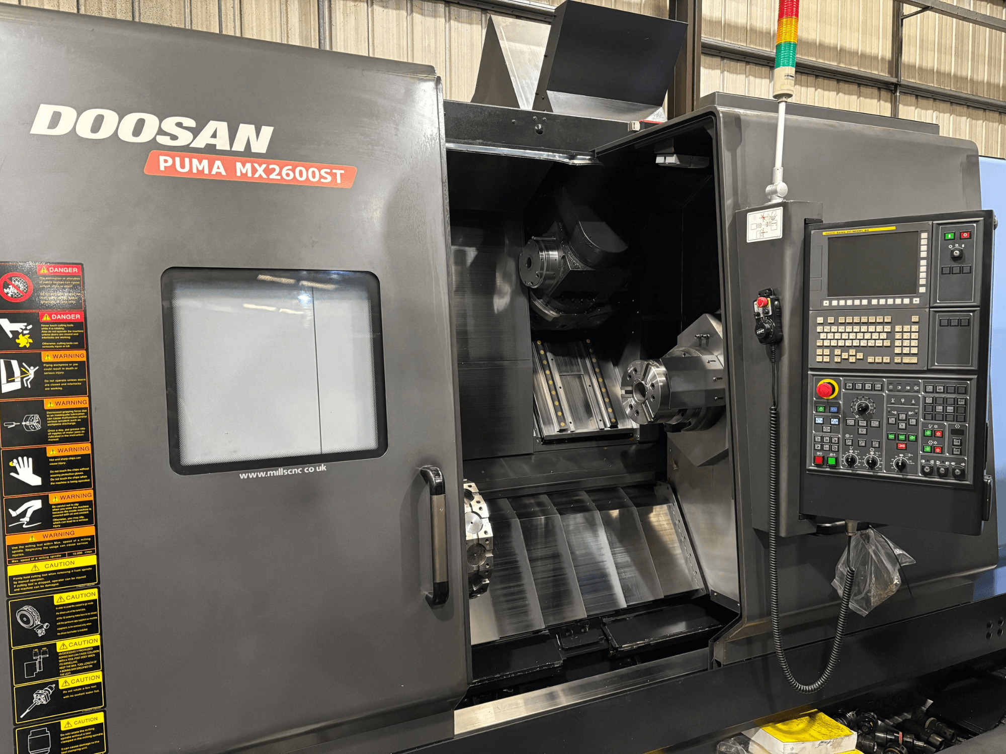 Torno CNC Doosan PUMA MX2600ST, vista frontal, mostrando el panel de control, el husillo y la zona de mecanizado con advertencias de seguridad visibles.