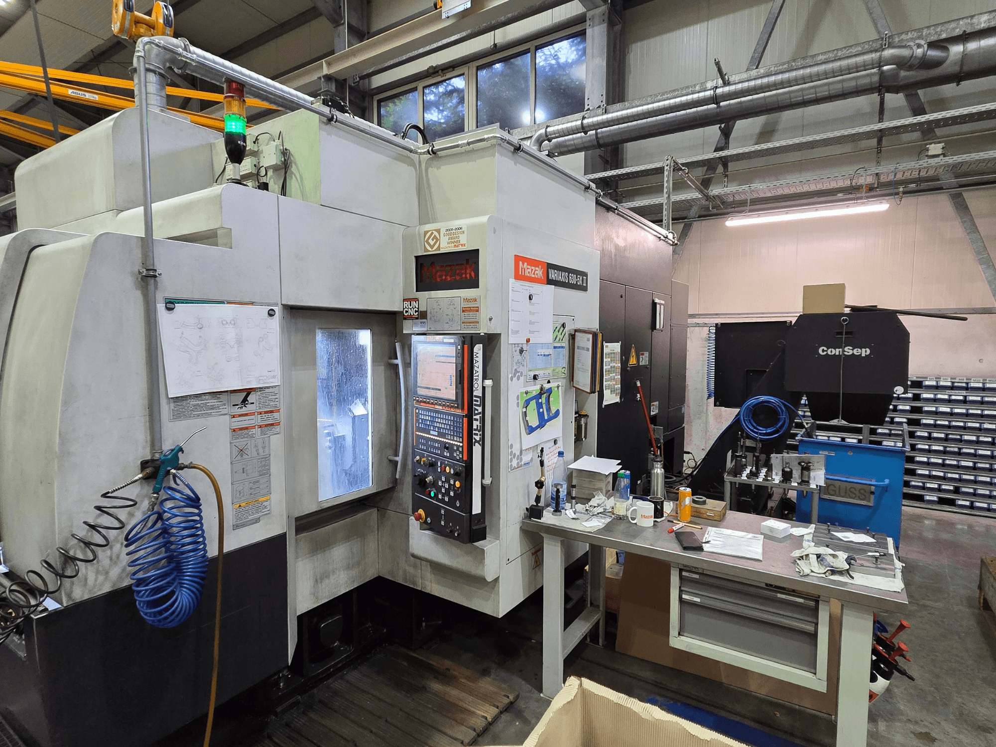Máquina CNC Mazak Variaxis 630-5X en un taller, vista frontal con panel de control, manguera y accesorios sobre mesa lateral.