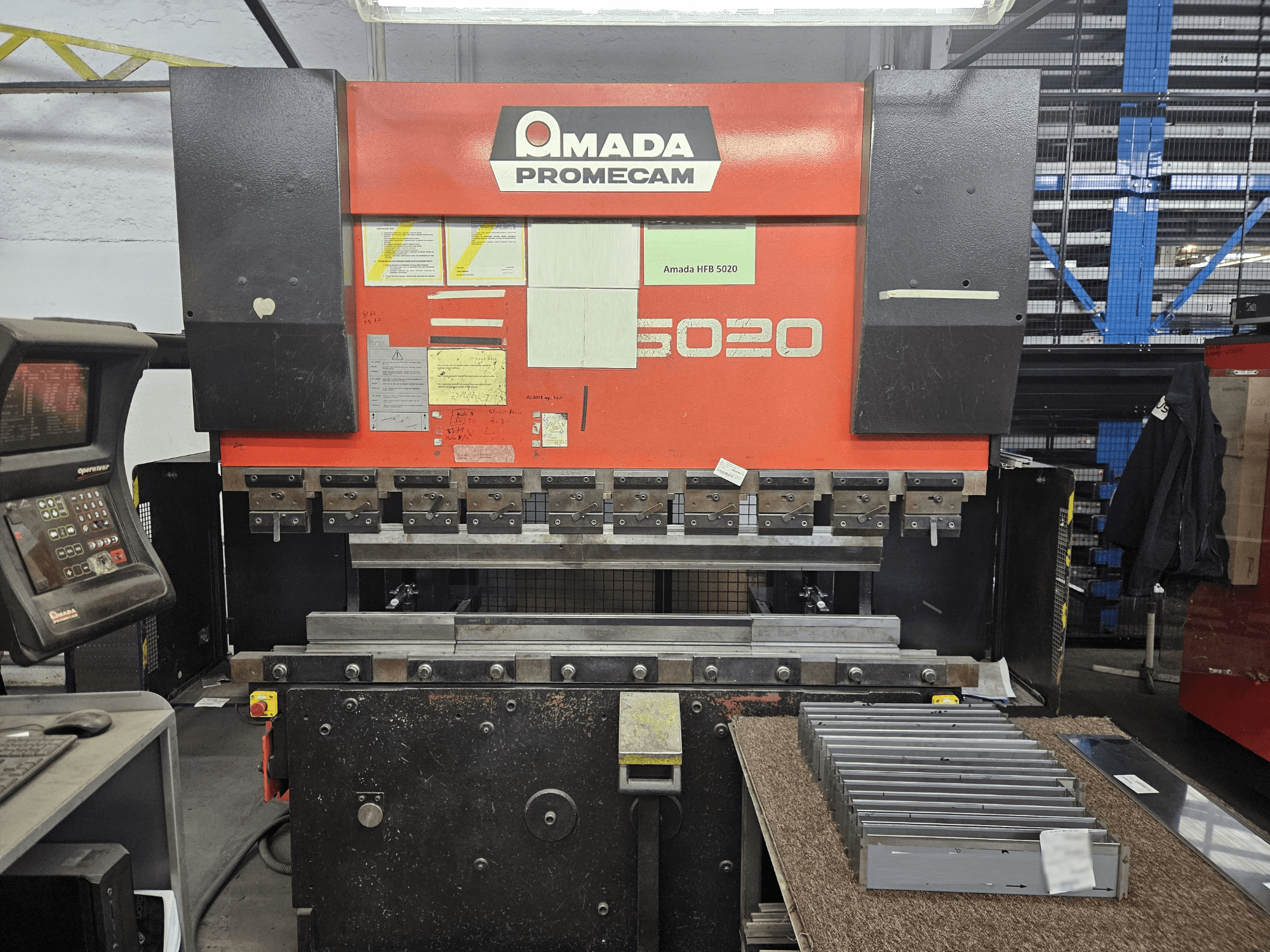 Plegadora Amada HFBO 5020 en vista frontal, con panel de control y piezas metálicas apiladas en la superficie de trabajo.