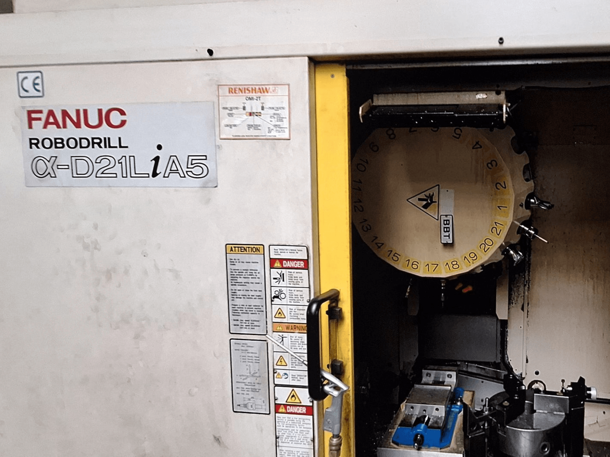 FANUC Robodrill α-D21LiA5, vista frontal. Muestra el carrusel de herramientas y la zona de mecanizado con pinzas y advertencias en la puerta.