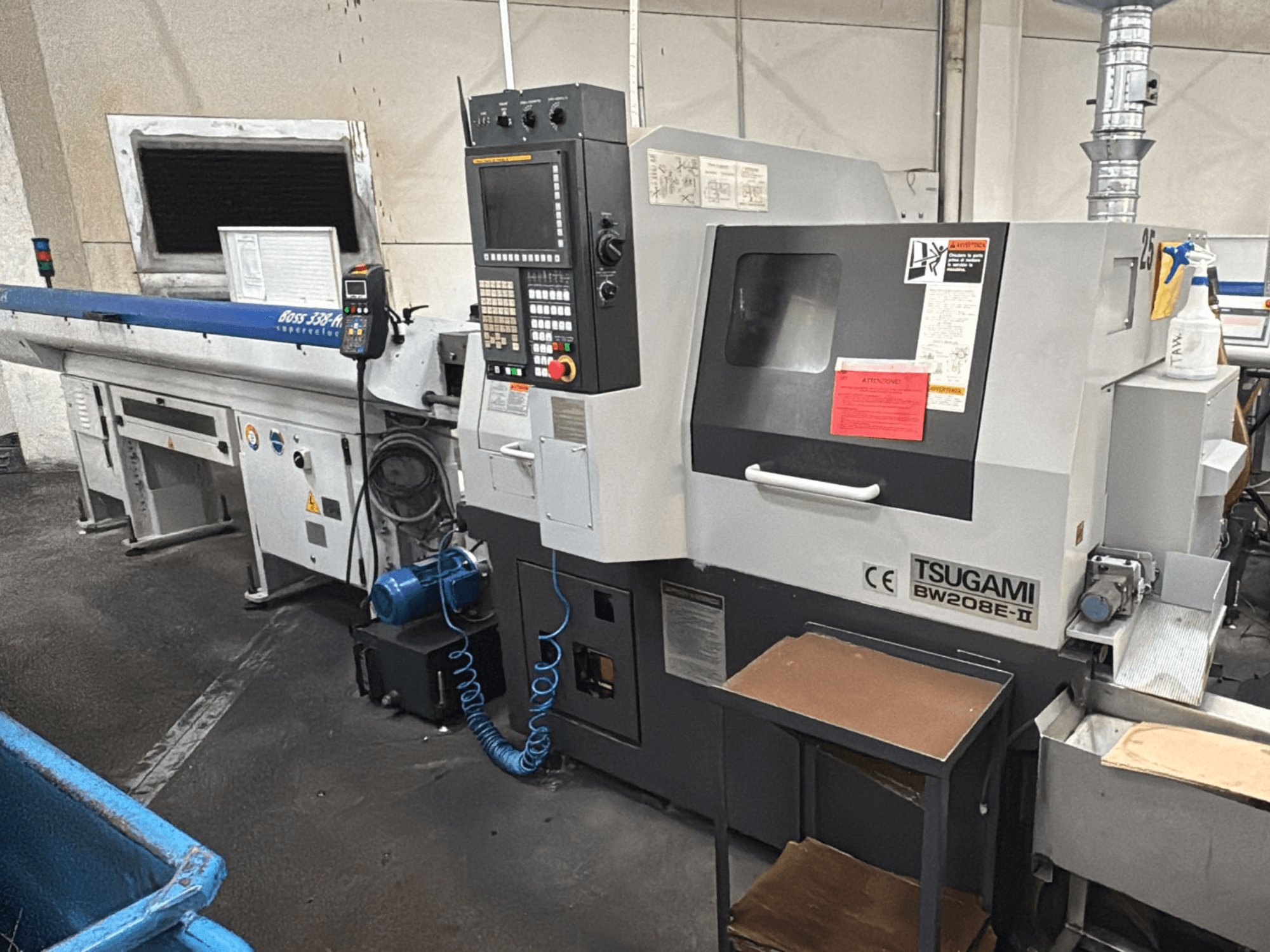 Máquina CNC Tsugami BW208E-II en un taller, mostrada desde un ángulo frontal, con el panel de control y las etiquetas de seguridad visibles.