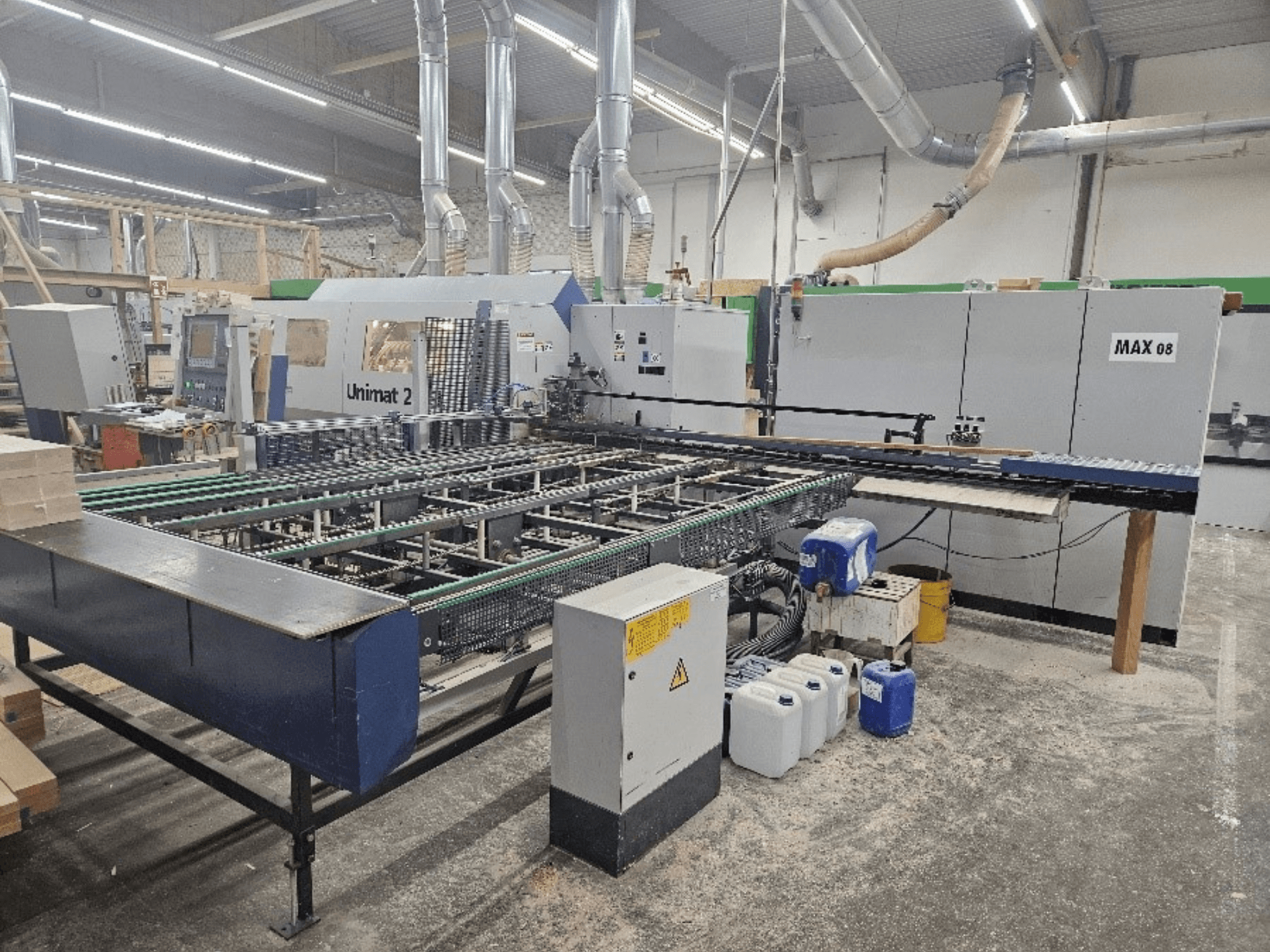 Vista lateral de una máquina CNC Unimat 2 en un taller, con cintas transportadoras y componentes técnicos con la unidad Max 08 visible.