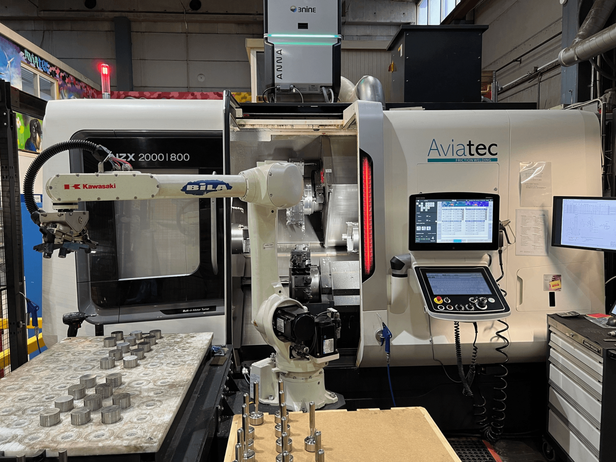 Torno horizontal DMG MORI NZX 2000/800SY con brazo robótico Kawasaki, panel de control y componentes metálicos sobre la mesa de trabajo.