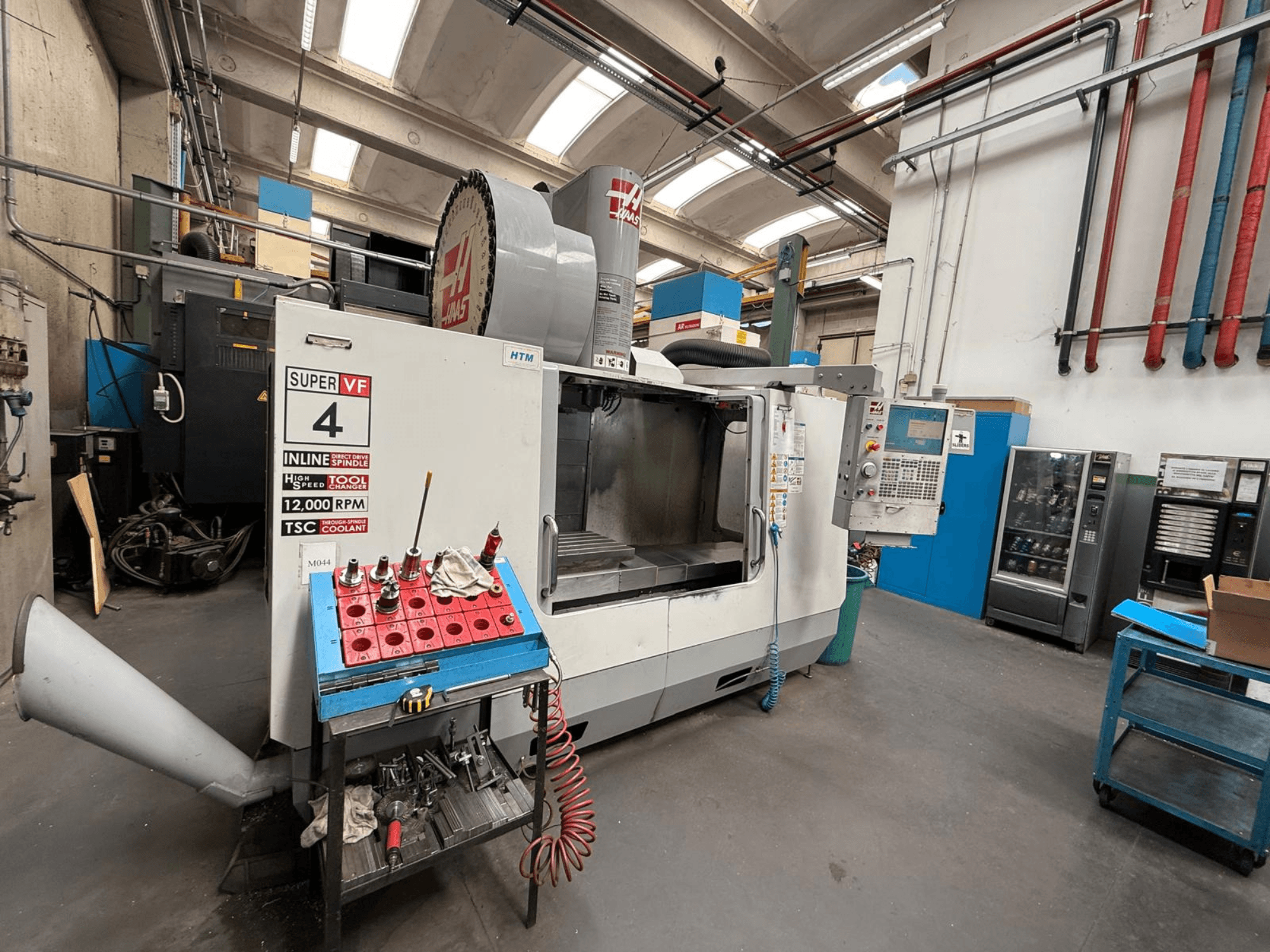 Fresadora CNC HAAS Super VF 4 con panel de control, husillo de 12.000 rpm y herramientas en un estante de almacenamiento azul, vista lateral.