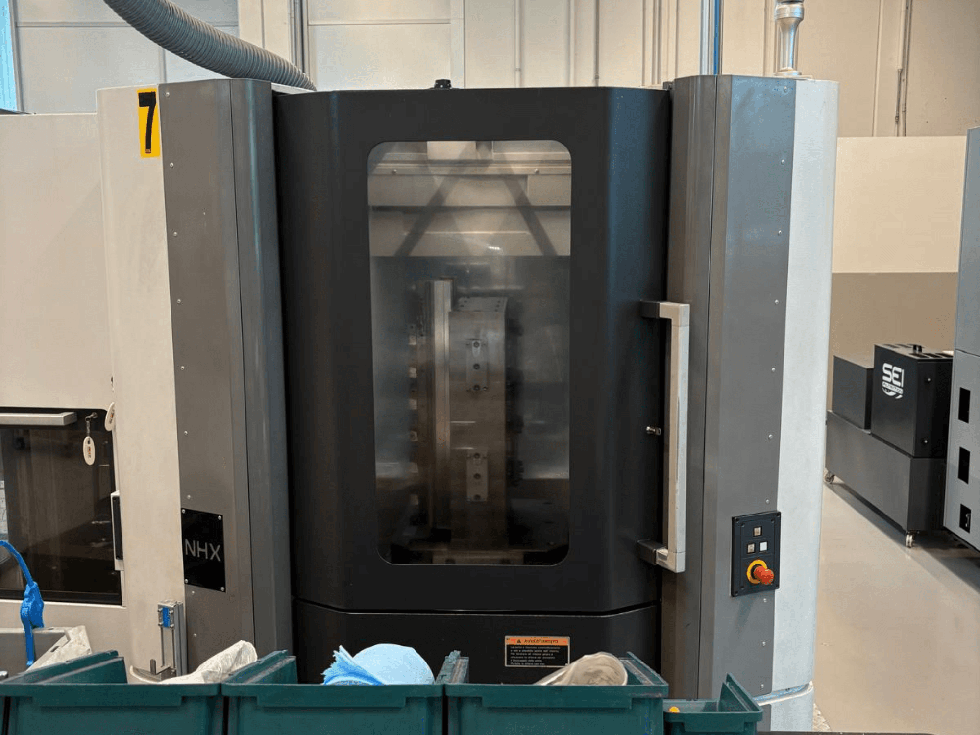 Máquina CNC NHX en vista frontal, mostrando el panel de control y la pantalla clara; situada en un taller con contenedores verdes cerca.