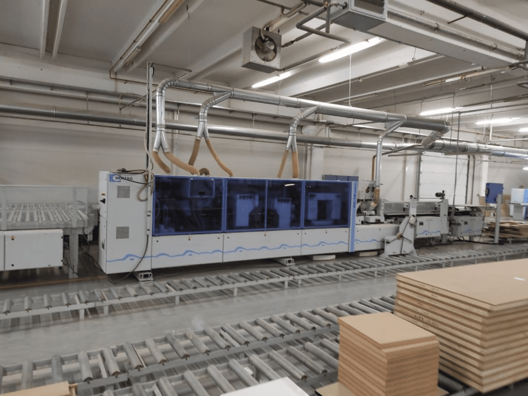 Máquina de producción HOMAG en una fábrica, vista frontal, con carcasa azul y sistema de transporte con planchas de contrachapado apiladas.