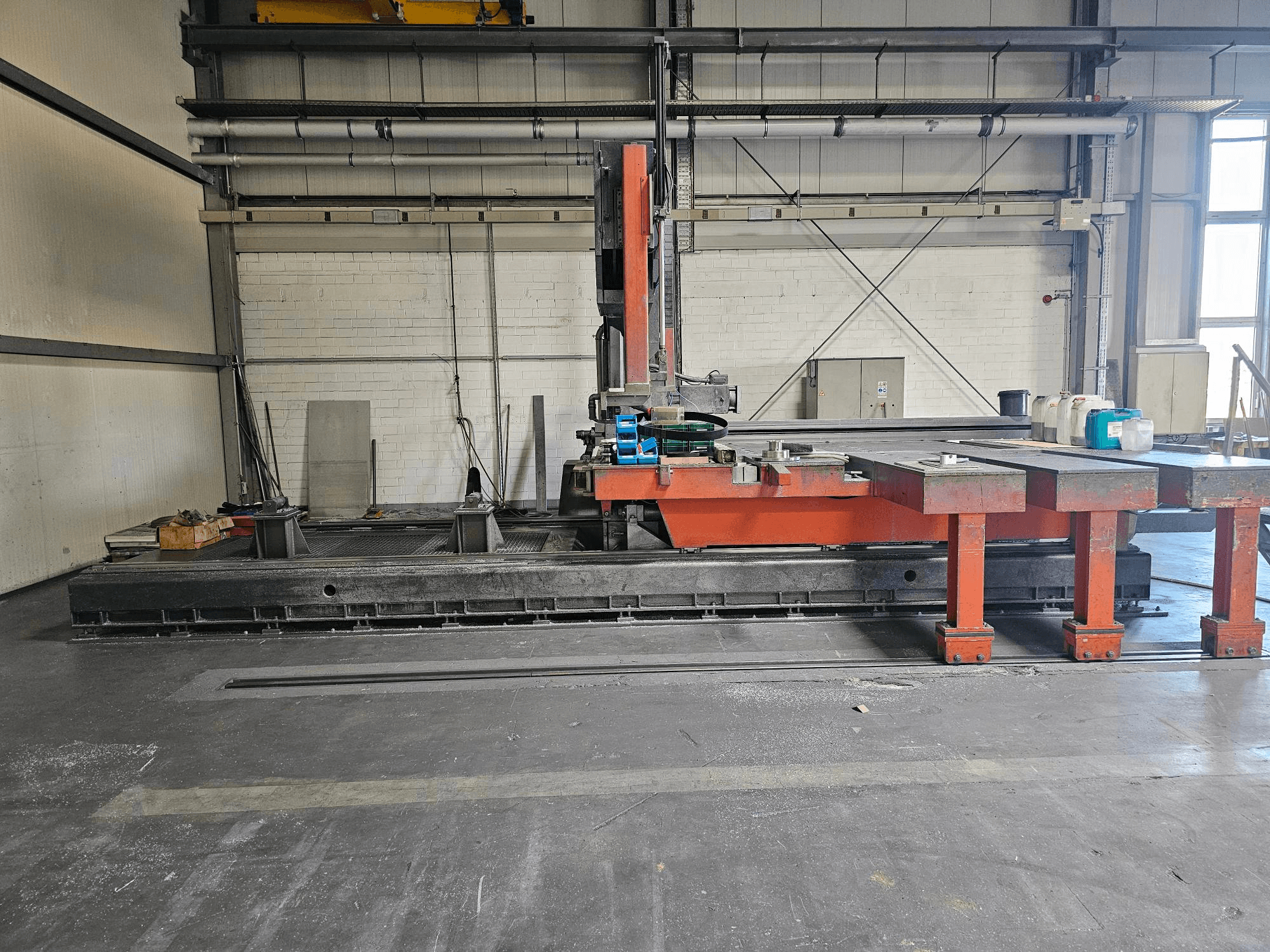 Máquina de corte industrial en un taller abierto, de color naranja y negro con una gran mesa; vista lateral que muestra los accesorios y el espacio de trabajo.