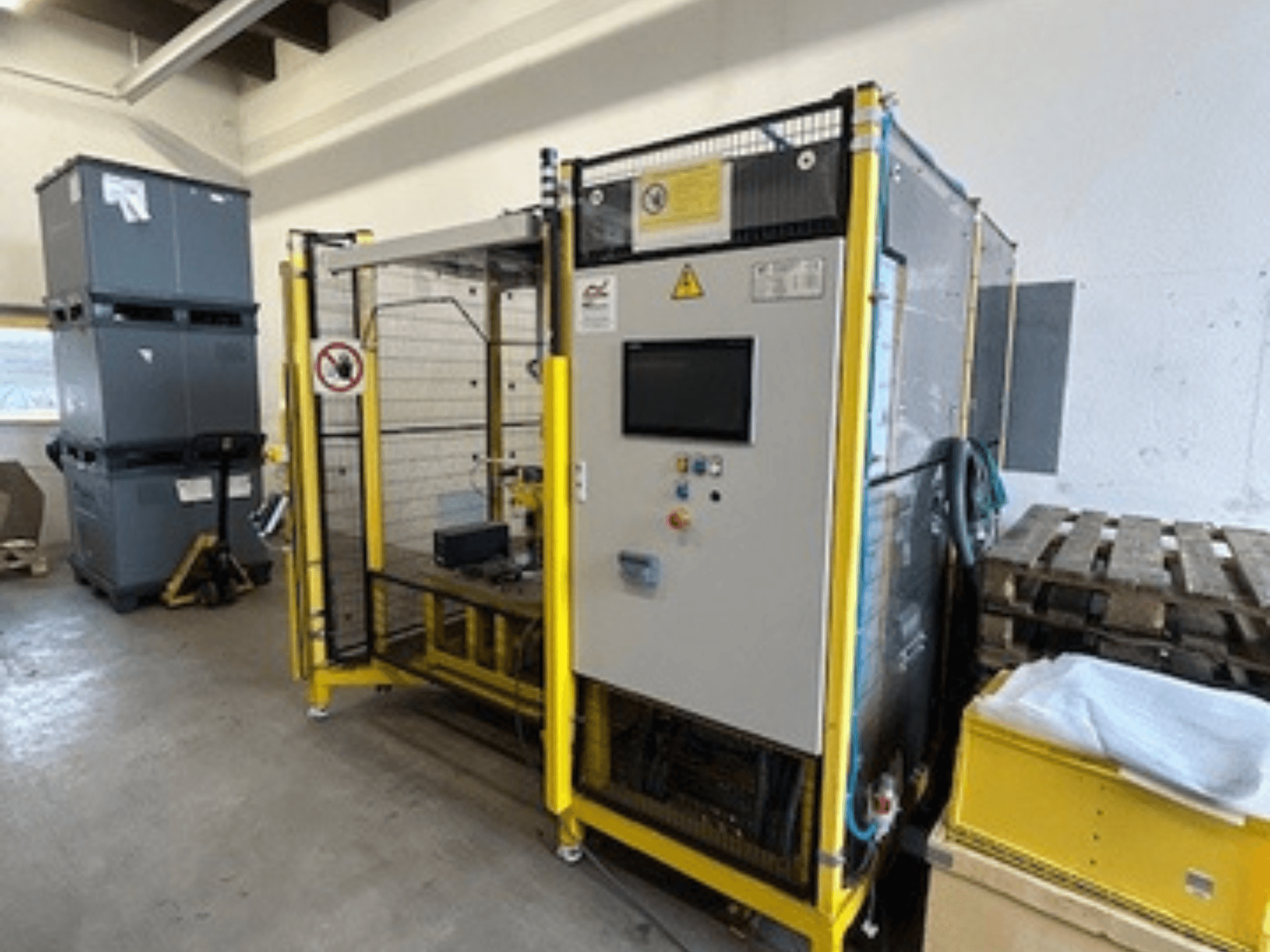 Máquina industrial con panel de control y dispositivos de seguridad, vista frontal; bastidor amarillo, contenedores grises y palés al fondo.