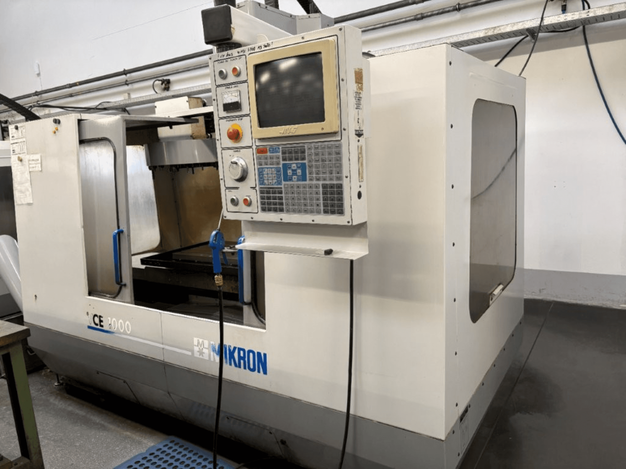 Máquina CNC Mikron CE 2000, vista frontal; presenta un panel de control con botones y pantalla, exterior plateado y zona de trabajo.