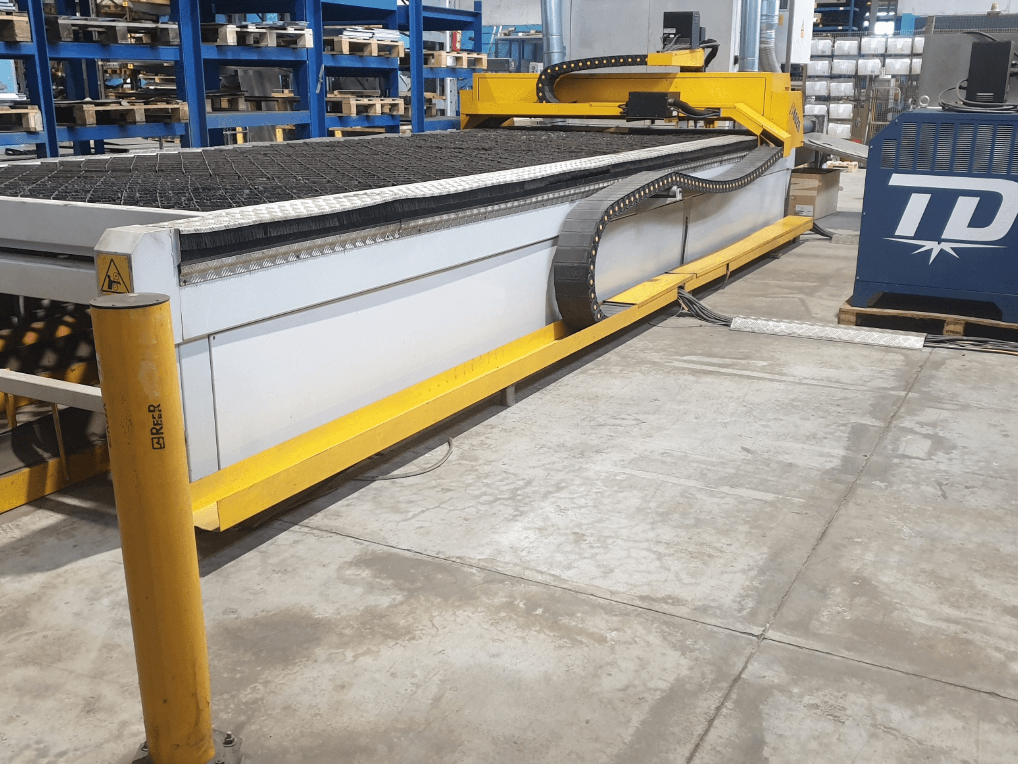 Máquina de corte industrial con embellecedor amarillo, vista frontal, con bancada texturizada y panel de control, colocada en la planta de una fábrica.