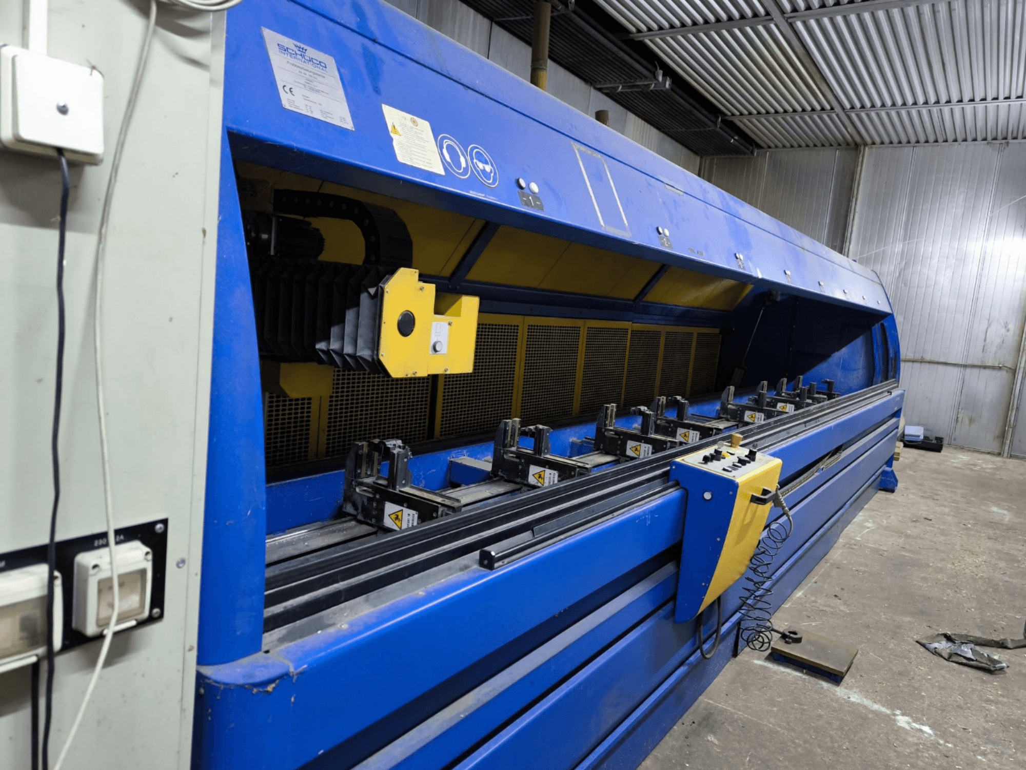 Máquina de corte industrial Schneider en azul, vista lateral, con panel de control, correas y múltiples estaciones de sujeción visibles.