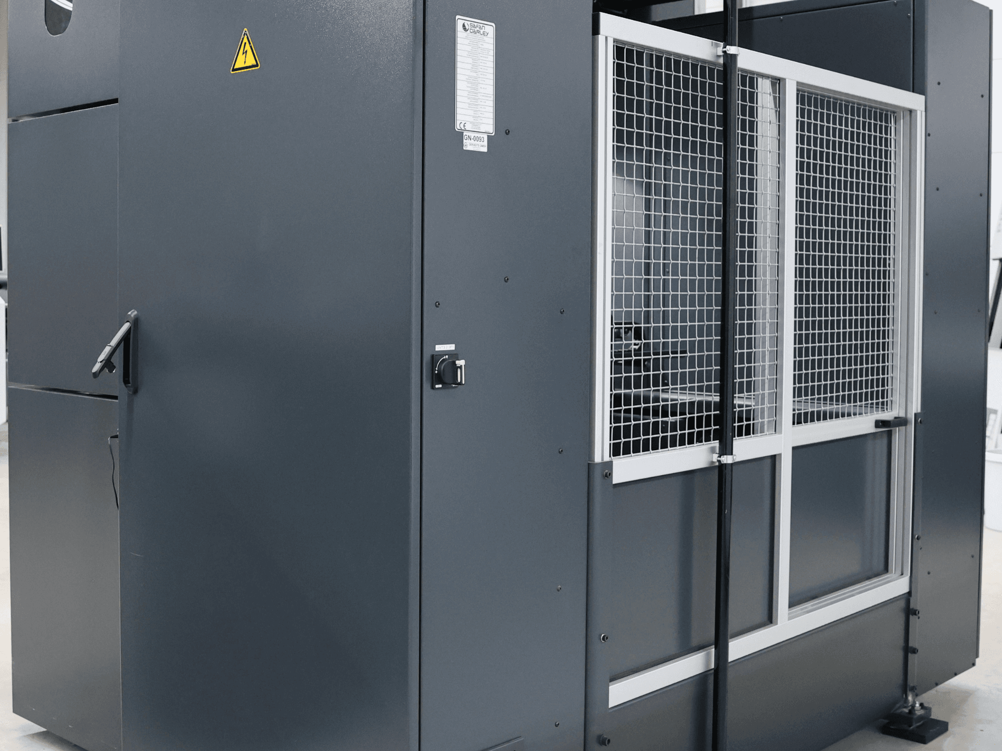 Máquina industrial gris con advertencia de seguridad, vista frontal mostrando puerta de malla y panel de control, modelo etiquetado.