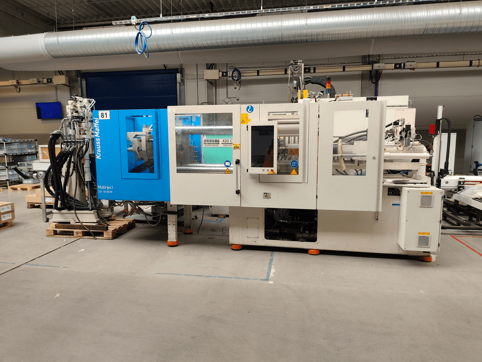 Máquina de moldeo por inyección hidráulica Krauss Maffei 110-180-180 CX V, vista lateral, en la que se aprecian el cuerpo azul y blanco, el panel de control y los conectores.