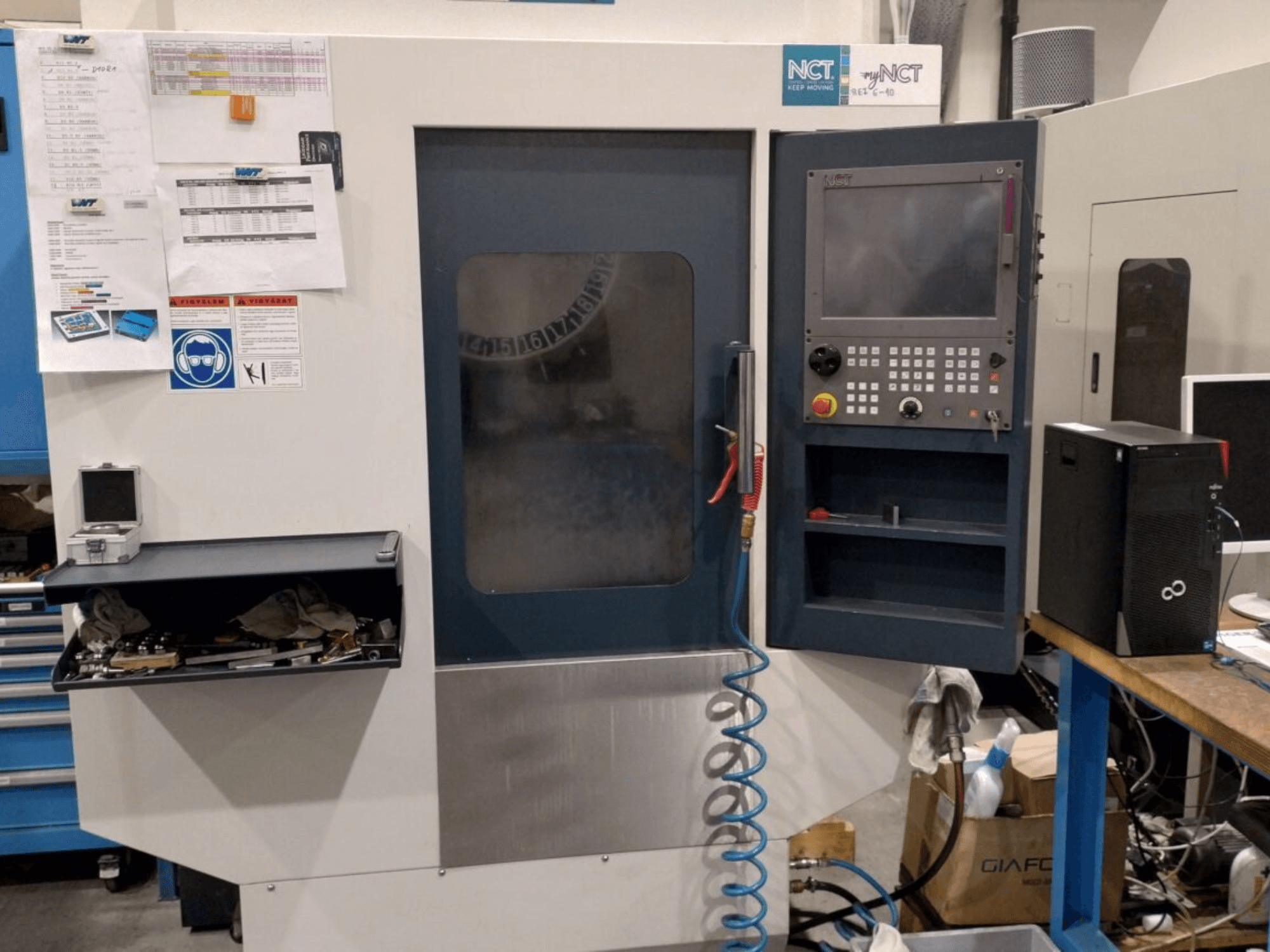 Máquina CNC NCT, vista frontal, presenta panel de control con teclado numérico, herramientas en cajón de almacenamiento y manual en la pared.