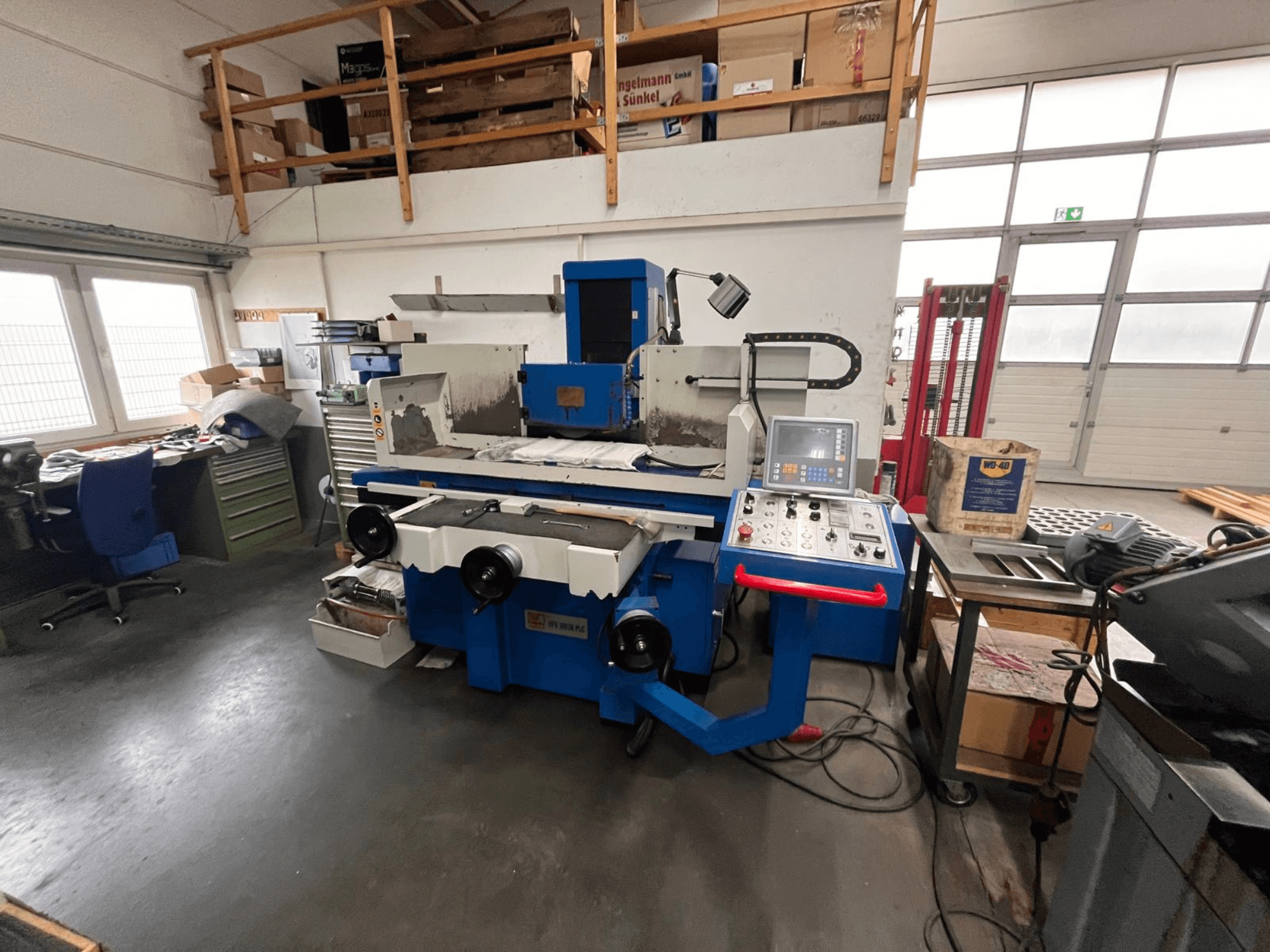 Amoladora de superficies azul con panel de control digital, vista frontal, mostrando el desorden del espacio de trabajo y un armario de herramientas verde.