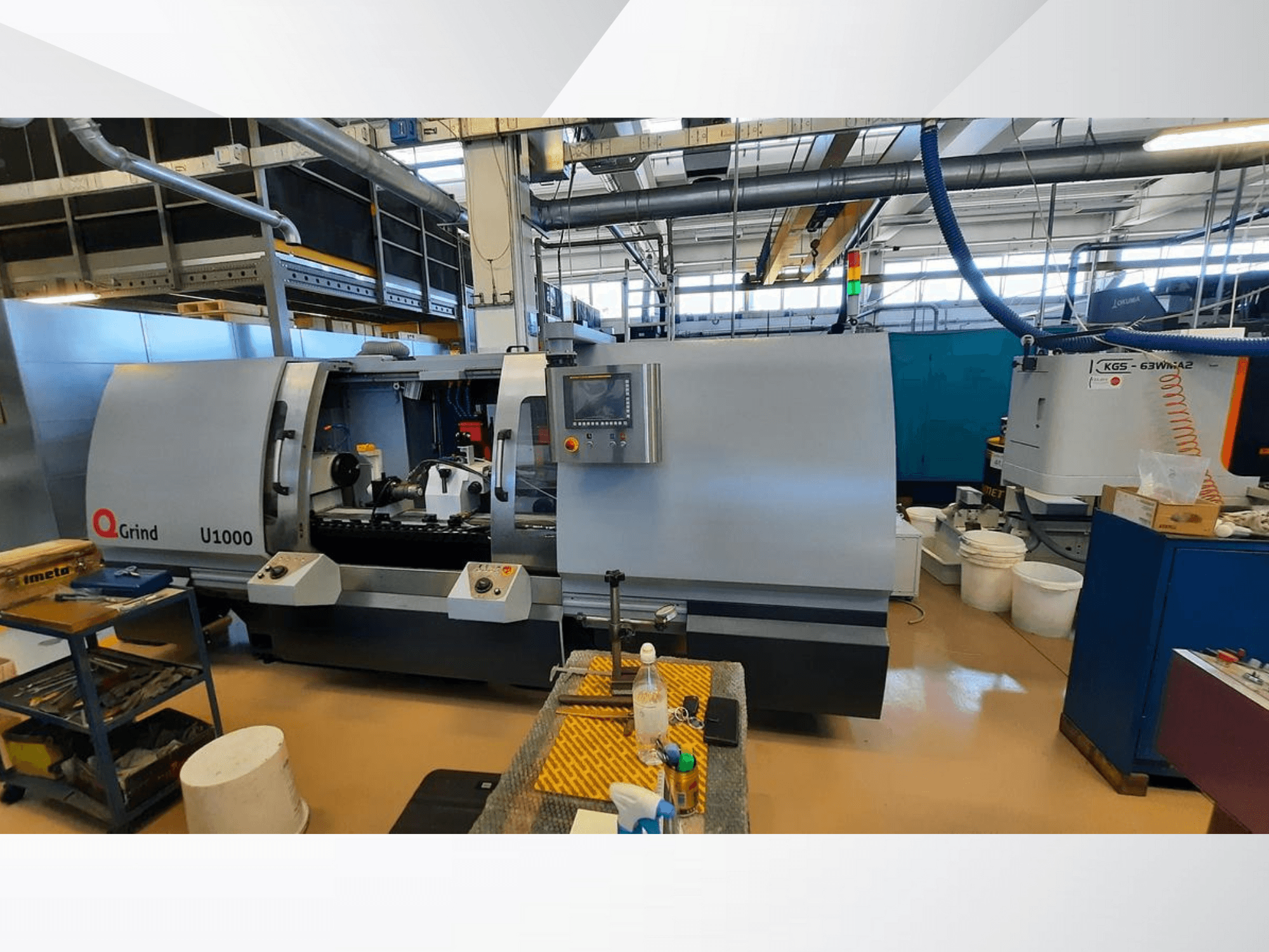 Máquina Q-Grind U1000 en un taller, mostrando una vista frontal con panel de control y zona de corte abierta, rodeada de equipos.