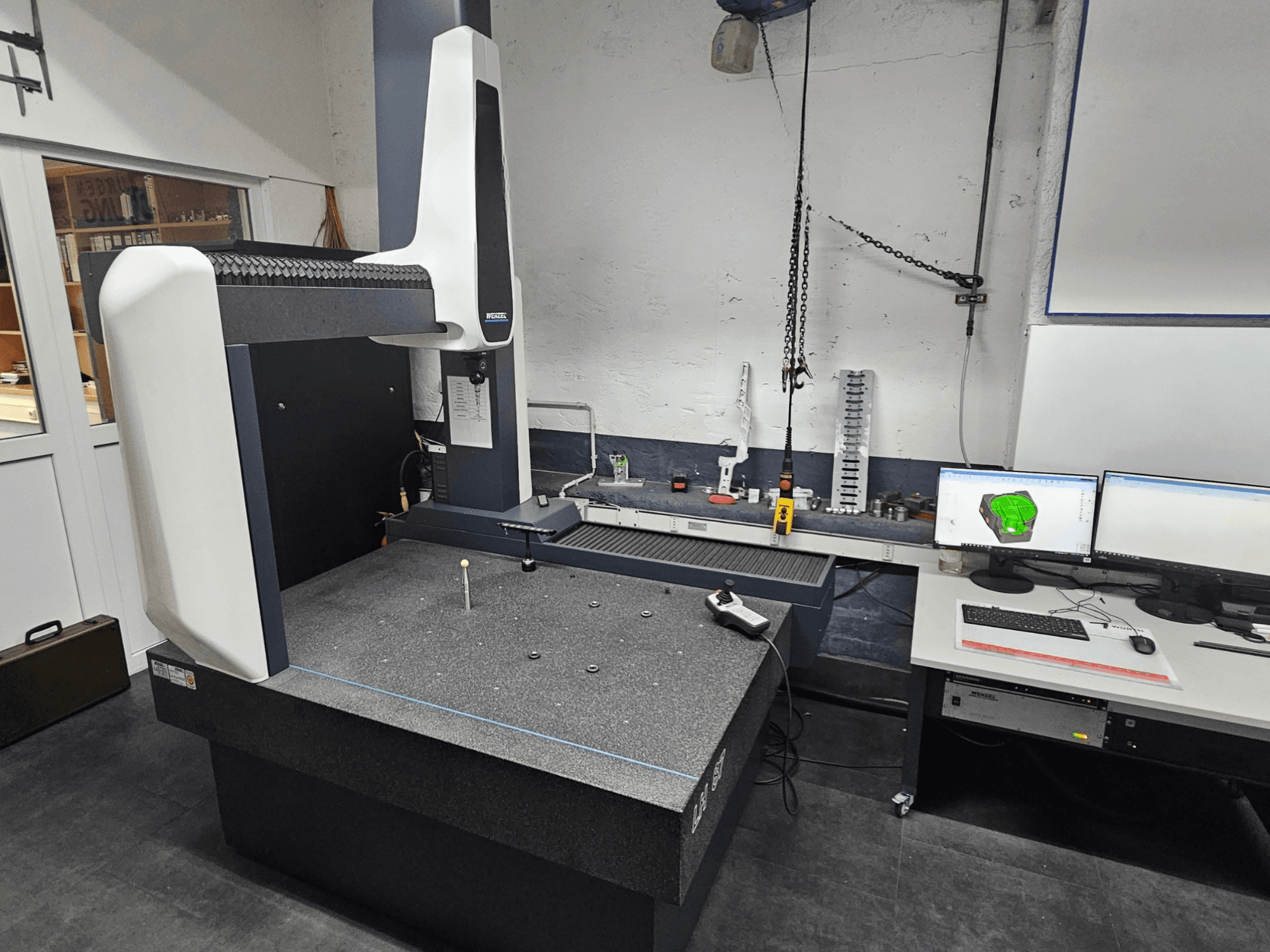 Máquina de medición por coordenadas Wenzel LH 87 en un taller, vista frontal, con una sonda en la mesa de granito y monitores dobles que muestran el diseño CAD.