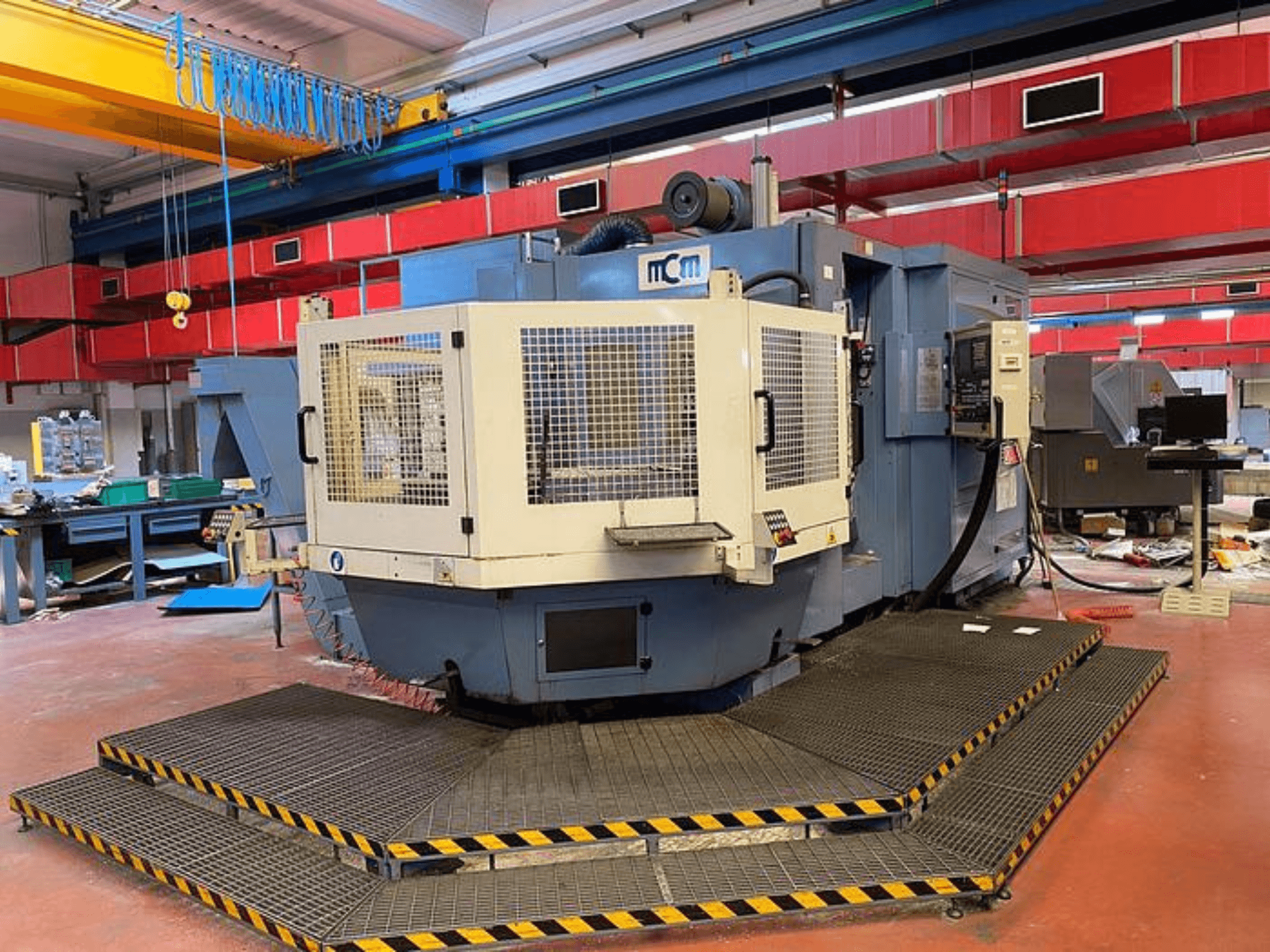 Máquina CNC MCM en una fábrica, vista frontal con jaula de protección, bordes de seguridad negros y amarillos, rodeada de equipos industriales.